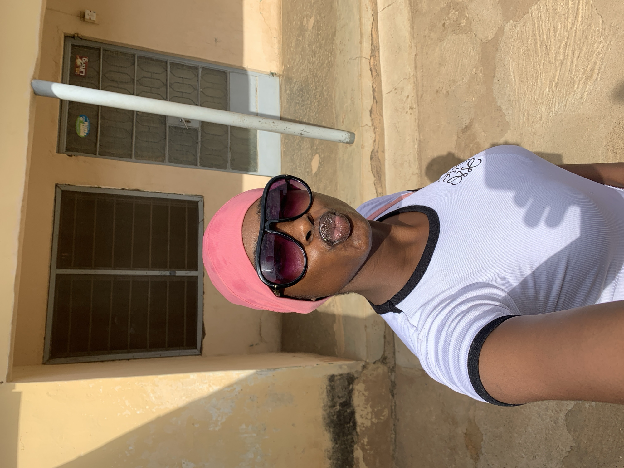 A woman with dark skin and pink headwear stands outside a building in Rafin Par, Nigeria, based on location metadata. She wears sunglasses and a white T-shirt with black trim. The building behind her has cream-colored walls and two windows, one above the other. Both windows are fitted with metal security bars or screens. A white pipe or conduit runs horizontally across the wall above the upper window. The ground or walkway in front of the building is a mix of concrete and dirt. The image appears to be taken in bright sunlight.