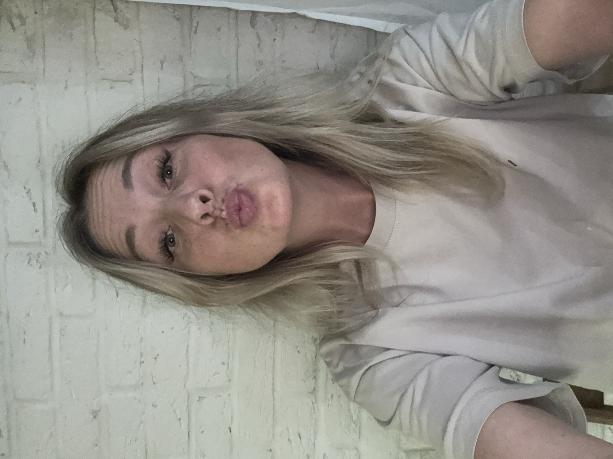 A woman with blonde hair and fair skin is positioned in front of a white brick wall. She is wearing a light-colored shirt. Her lips are pursed in a kissing gesture. The environment appears to be indoors. The city of Chaikovsky, Russia, is not discernable from the provided visual information.