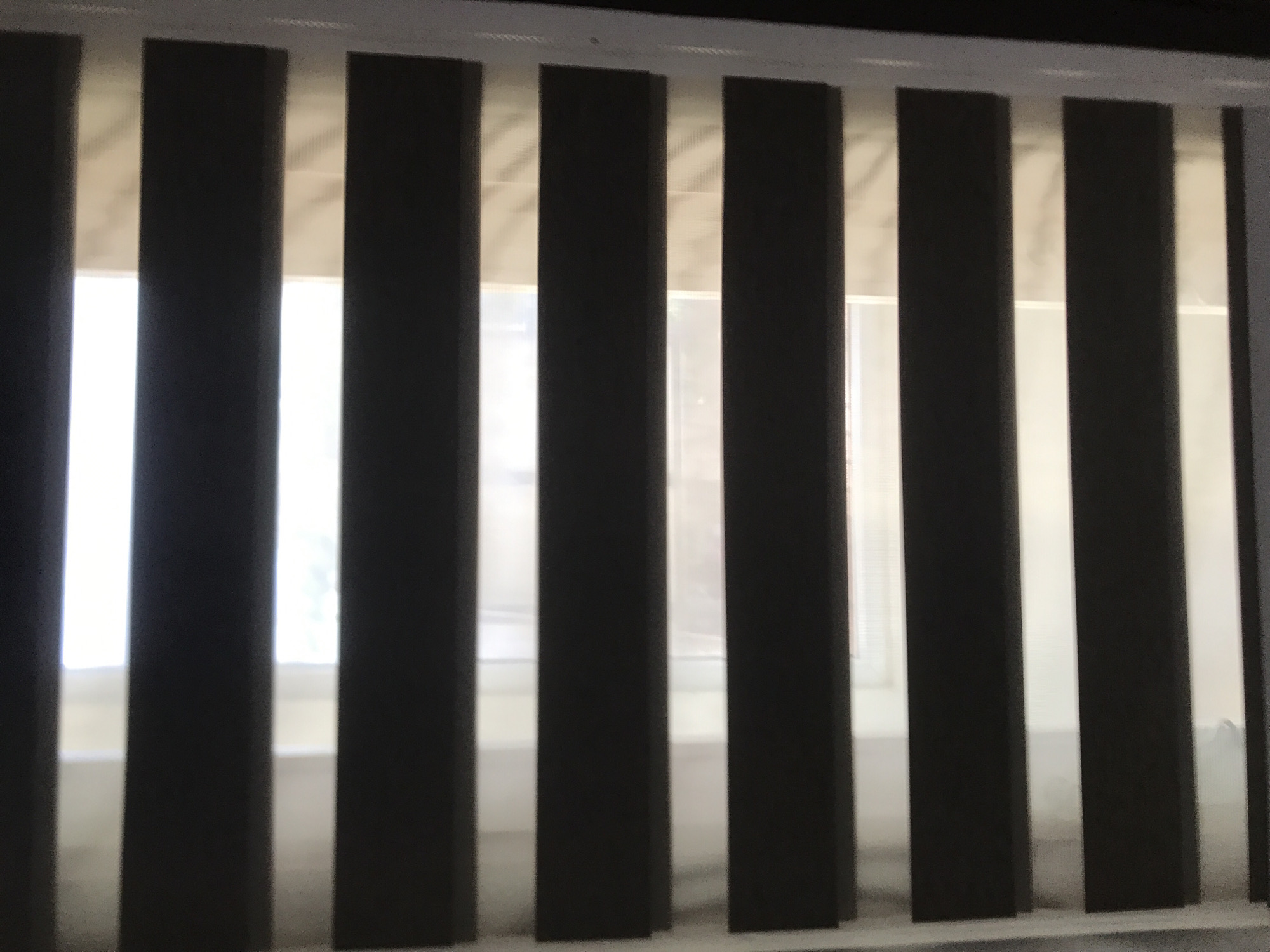 The image shows a close-up view of vertical blinds covering a window. The blinds are mostly closed, allowing some light to filter through. The location is unknown as the image provides no contextual clues to identify Banda Aceh, Indonesia. There are no people, subjects, objects, actions, or interactions explicitly visible in this image, only the blinds and the light coming through them.
