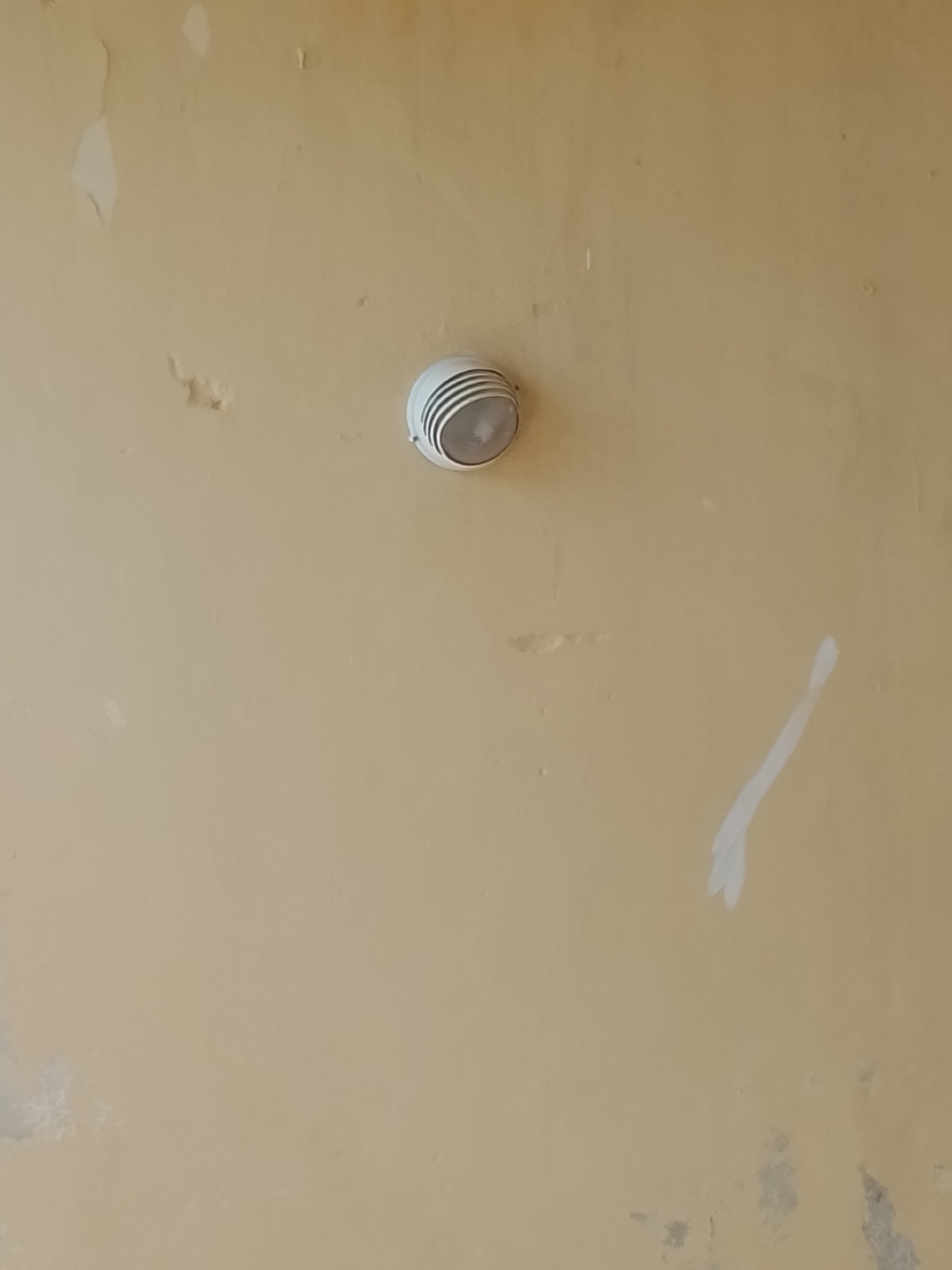 The image shows a close-up of a beige, textured wall with signs of wear and tear, including peeling paint and some water damage. A white circular light fixture, possibly a smoke detector or a small ceiling lamp, is mounted on the wall. The lighting suggests an indoor setting, possibly a public space or a room with an overhead light source. There are no people visible in the image. The overall impression is of a utilitarian, possibly older, interior wall. The location in Jalingo, Nigeria, is inferred from external knowledge, but the image itself does not contain any specific markers to identify the city.