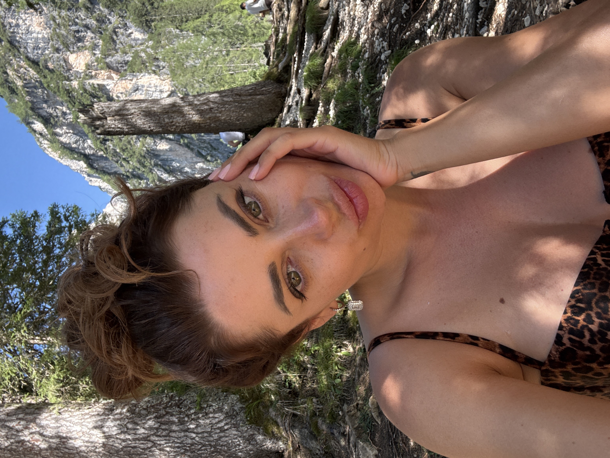 An overhead, close-up selfie shows a woman with her hair in a messy bun, resting her left hand on her cheek. She is wearing a leopard-print bikini top. She is positioned in the foreground, with a backdrop of trees and a steep, rocky mountain slope under a clear blue sky. The ground is covered with dirt, rocks, and roots. In the middle ground, two men are visible in the distance, one sitting and the other standing on a path. The overall scene suggests an outdoor setting, possibly a hiking trail in the mountainous region of Prags - Braies, Italy.