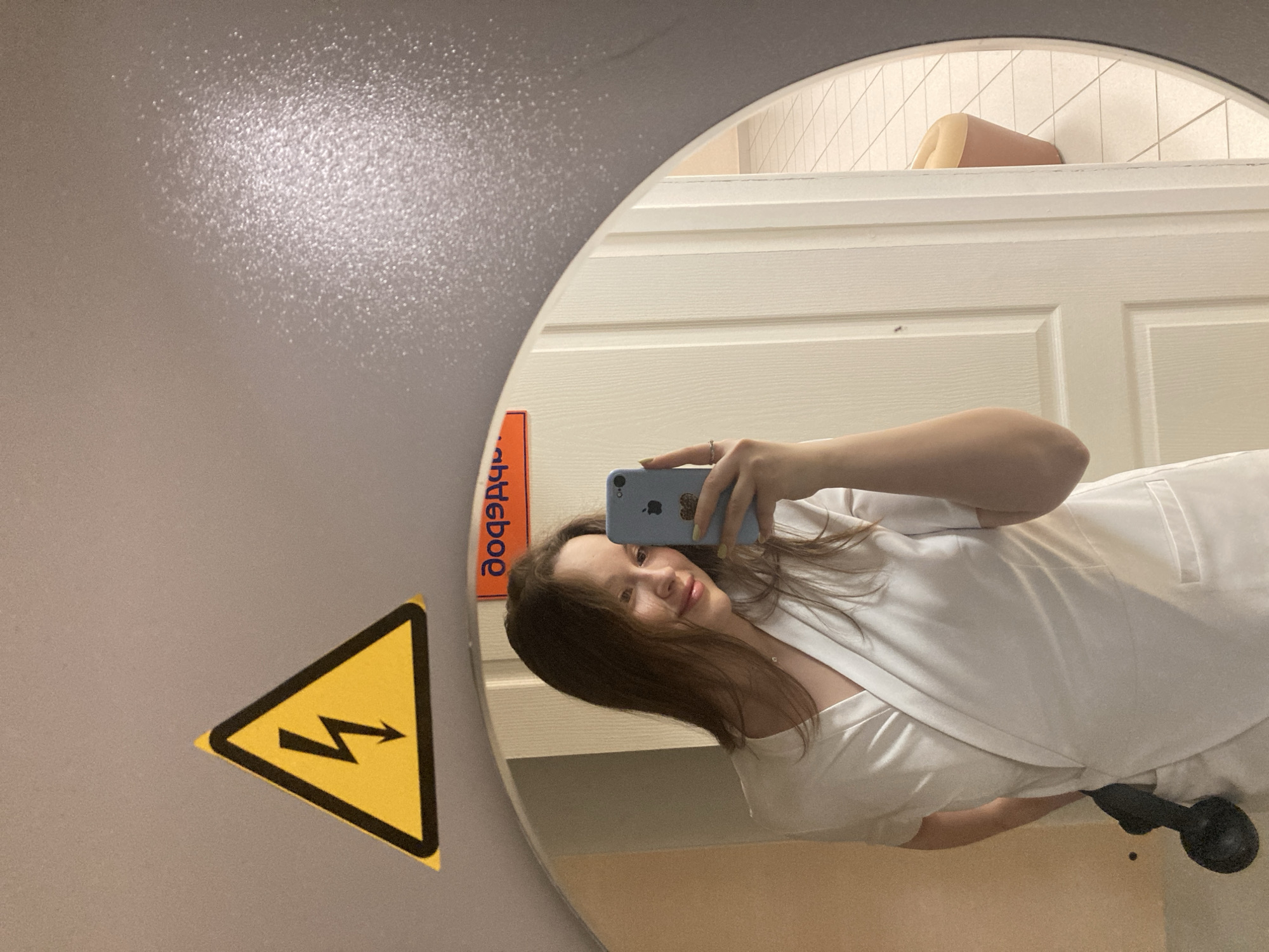 A woman is taking a mirror selfie in what appears to be a public restroom. The mirror is oval-shaped and reflects a white-tiled wall with beige accents above the door, and the woman holding a light blue iPhone. She is wearing a white outfit with a visible black object at the bottom, possibly a bag or item of clothing. A bright yellow hazard sign is visible on the wall outside the mirror. The sign has a black lightning bolt symbol inside a triangle. Above the mirror is an orange sticker or label with blurred text in Russian, likely indicating the location as Chaikovsky, Russia.