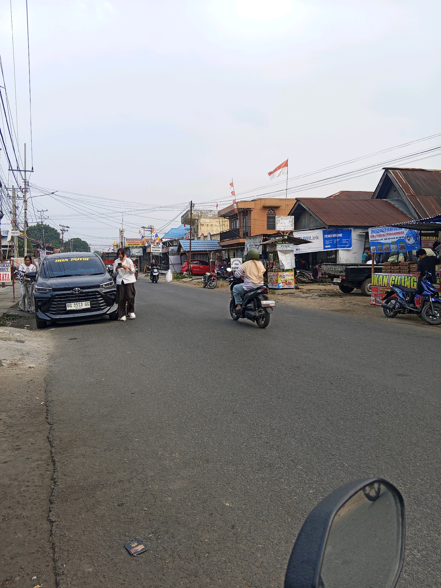 A street scene in Lubuklinggau, Indonesia, features a paved road with several parked and moving vehicles, including a black Toyota Avanza with a license plate reading "BG 1513 GG," a black Mitsubishi pickup truck, several motorcycles, and a red car. Two women are standing near the black Toyota Avanza, one holding a phone and the other looking at her phone. A person wearing a green helmet is riding a motorcycle away from the camera. Buildings line the right side of the street, including shops with signs, some displaying the Indonesian flag. Power lines and utility poles are visible in the background. The sky is overcast.