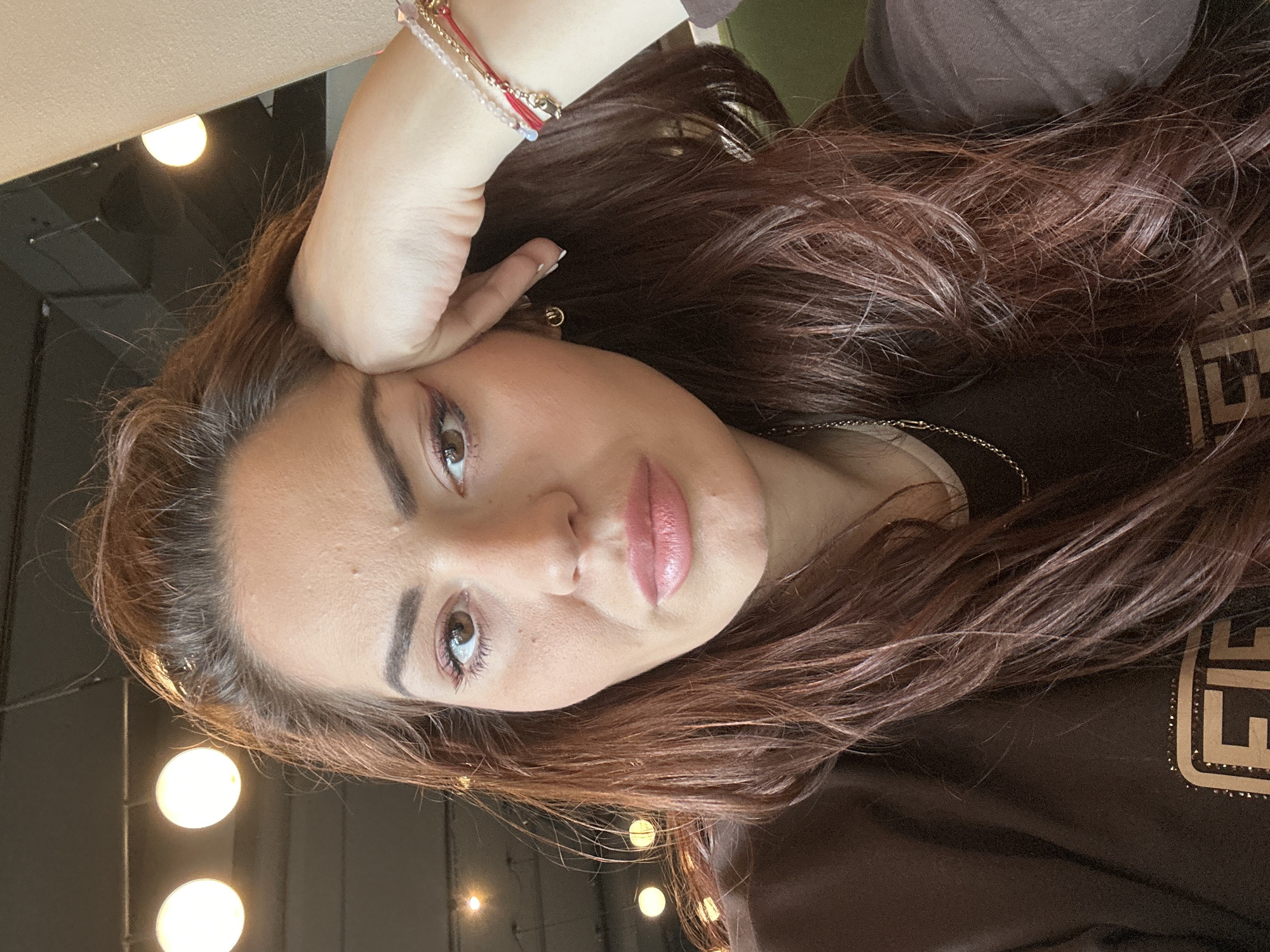 A medium-shot, angled selfie depicts a woman with light skin, light brown eyes, and auburn hair. She is wearing a brown top, and a bracelet is visible on her wrist. The background features multiple light fixtures, and what appears to be an indoor setting. The image gives no specific details to indicate the location being Wrocław, Poland.
