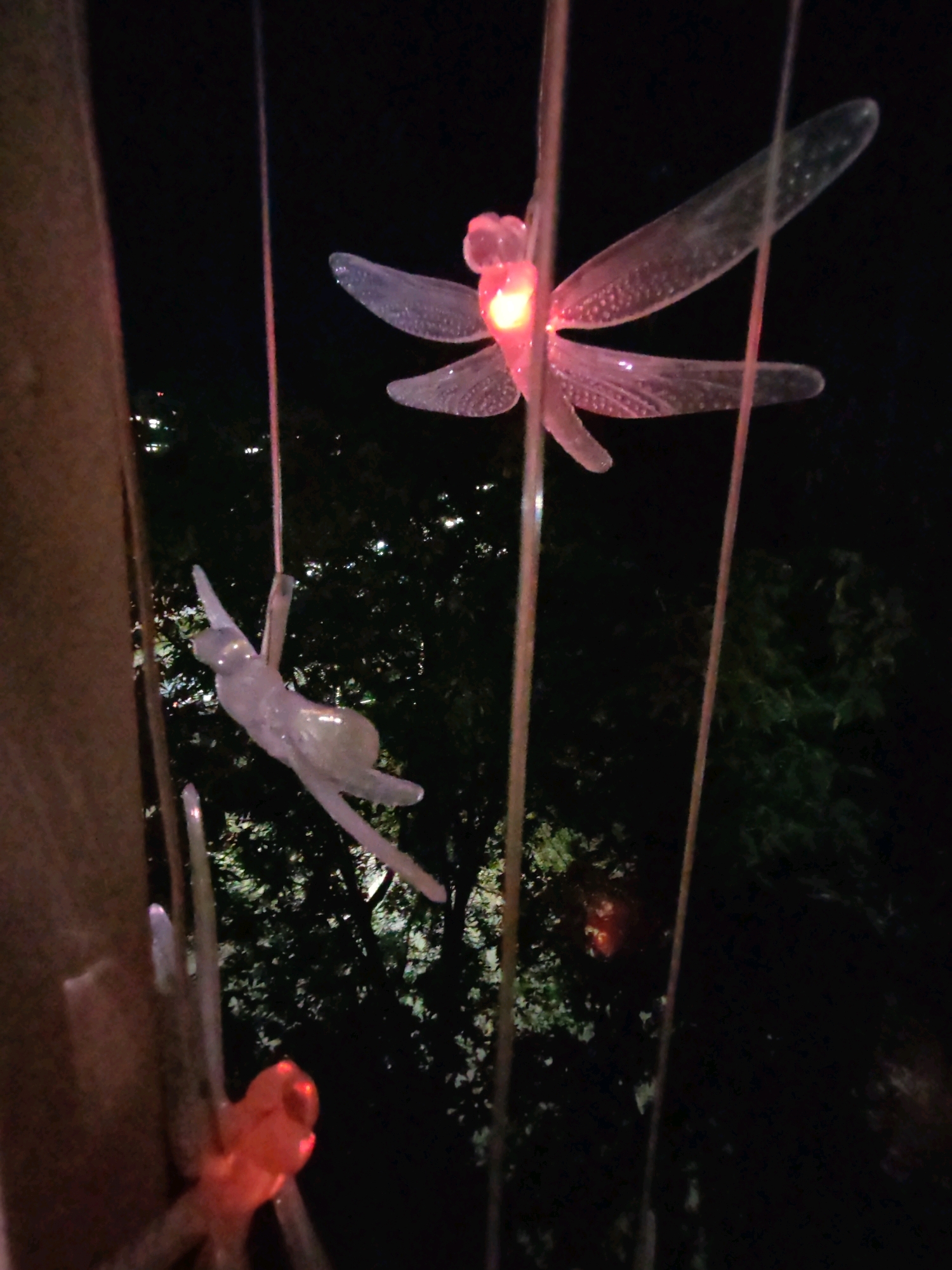 This is a nighttime photograph taken outdoors in Izhevsk, Russia. The foreground features several thin, transparent poles with lights at their ends. One pole has a brightly lit, pink dragonfly-shaped ornament hanging from it, with its wings spread. Another pole has a pale, translucent ornament resembling a leaping cat. In the background, there are dark trees with some scattered lights, possibly from buildings or streetlights, indicating an urban or park setting within Izhevsk. The overall scene suggests an outdoor decorative lighting installation at night.