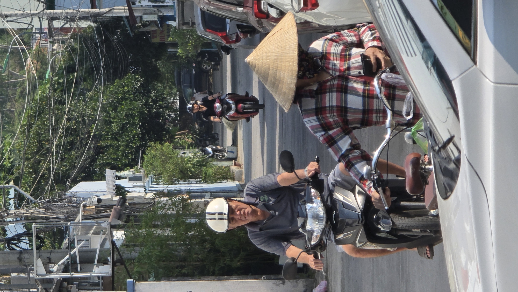 The image depicts a street scene in Bắc Nha Trang, Vietnam. A man wearing a white helmet and a grey shirt rides a scooter in the foreground. Beside him is a person in a red plaid shirt and a traditional Vietnamese conical hat, also on a scooter, situated next to a parked white car. In the background, another person on a red scooter is visible, riding away from the camera. The environment includes buildings, power lines, trees, and other parked vehicles lining the street.