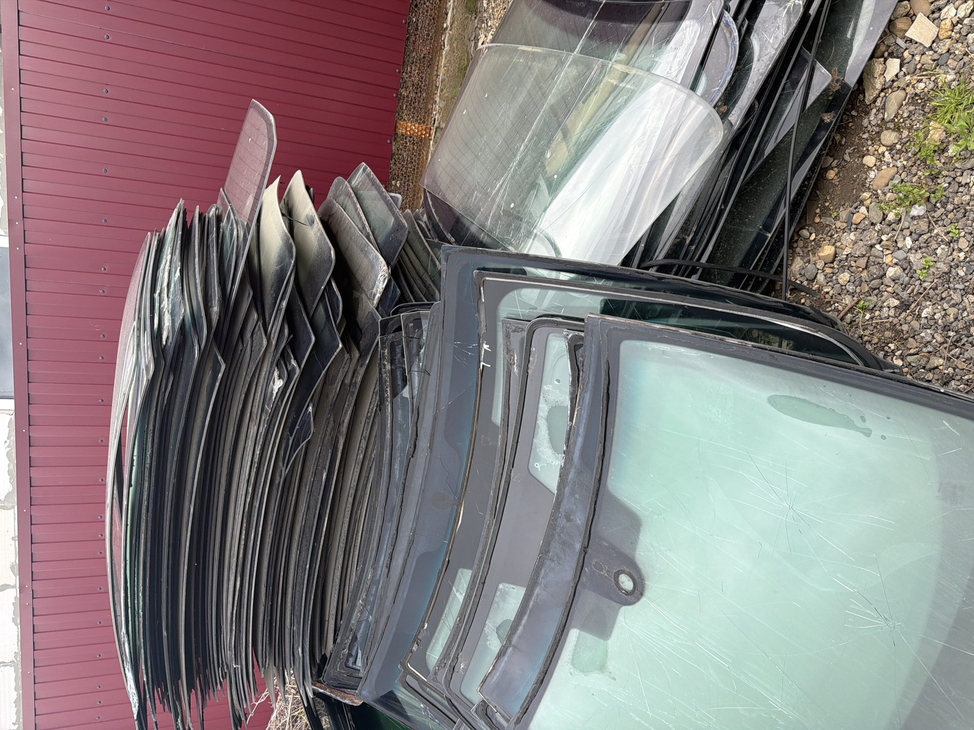 The image shows a large collection of stacked windshields, likely from automobiles, leaning against a reddish-brown corrugated metal wall. The setting appears to be outdoors, on gravel ground. Some of the windshields exhibit cracks and damage. The location appears to be an automobile scrapyard or similar storage area.