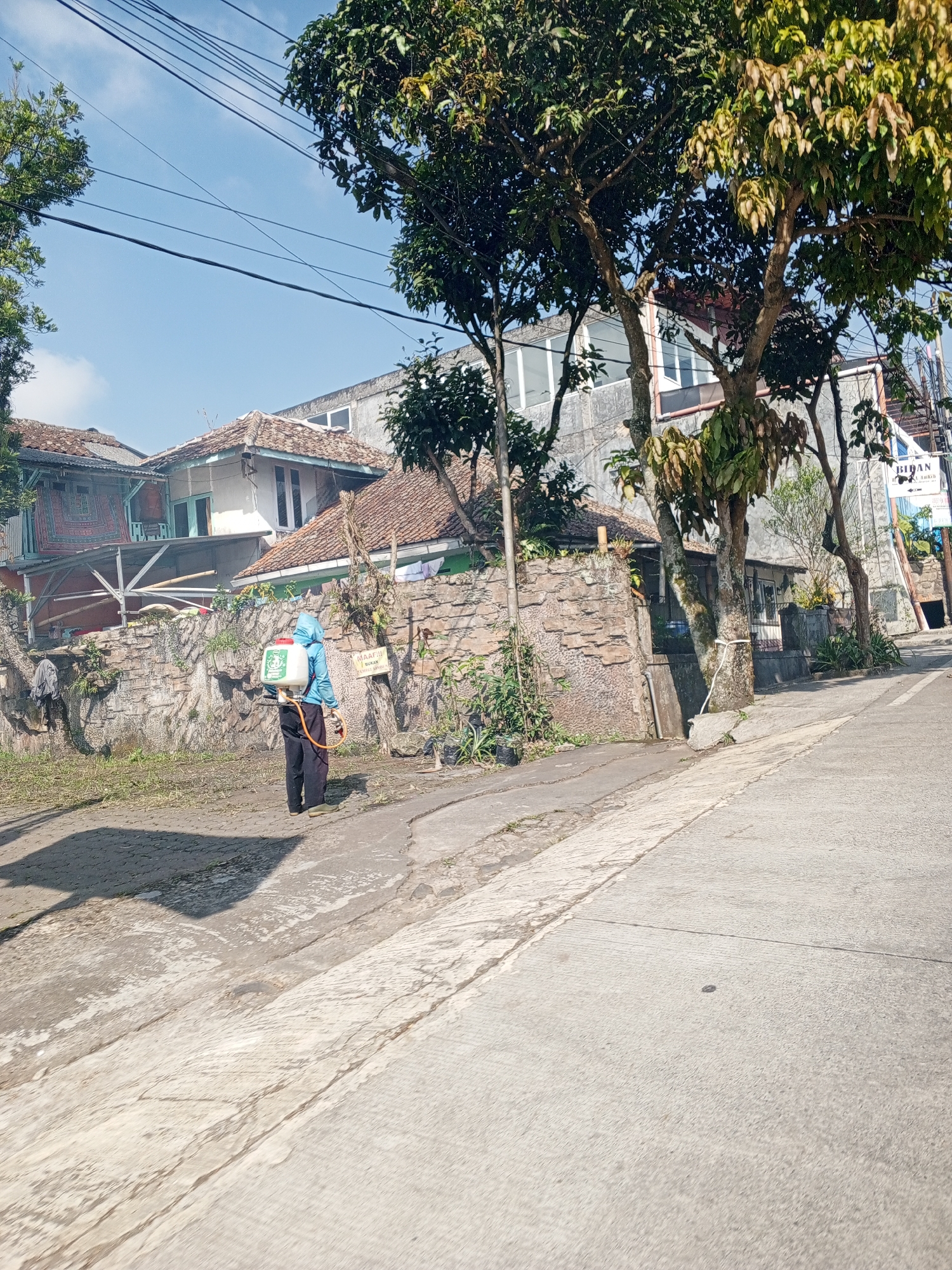 This detailed image captures an outdoor scene on a clear, sunny day in Weddagala, Sri Lanka. The perspective is from a paved road or driveway, looking towards a residential area. In the mid-ground, a single individual is visible, positioned with their back mostly to the viewer, facing towards a stone wall and various residential structures. This person is wearing a light blue or teal hooded jacket or protective suit and dark trousers. They are equipped with a white and red backpack sprayer, from which an orange hose extends downwards. Their posture suggests they are either preparing to spray, actively spraying, or have just completed a spraying action, possibly involving pest control, weed management, or disinfection. To the left of the individual, the ground consists of a concrete sidewalk transitioning to an unpaved area covered with sparse grass and earth. Behind the individual and extending across the left-mid portion of the image is a roughly constructed stone wall, appearing to form a boundary for the properties beyond. Affixed to this stone wall, slightly to the right of the person, is a small rectangular white sign with black text, partially legible, reading "MAAF!!!" on the top line, and "INI BUKAN JALAN UMUM" below it, which translates to "SORRY!!! THIS IS NOT A PUBLIC ROAD." Behind the stone wall, several buildings are visible. To the far left, a house with a reddish-brown tiled roof and light-colored walls is partially obscured by trees. A piece of patterned red and white fabric or a rug hangs over a railing on a structure in front of this house. Another house with a similar tiled roof and light-colored walls, featuring white window frames, stands directly behind the sprayer. Further to the right and slightly behind these structures is a taller, more modern multi-story building with a flat roof and large rectangular windows. In the far right background, another building with a sign that partially reads "BIDAN" (likely indicating a midwife clinic or similar facility) is visible. Numerous trees with dense green foliage are scattered throughout the scene, some framing the clear blue sky, while others grow between and in front of the buildings. Sunlight is bright and direct, casting distinct shadows from the trees onto the road and sidewalk, as well as a shadow from the person and their equipment onto the ground to their left. Overhead, several black utility lines (likely power or communication cables) crisscross