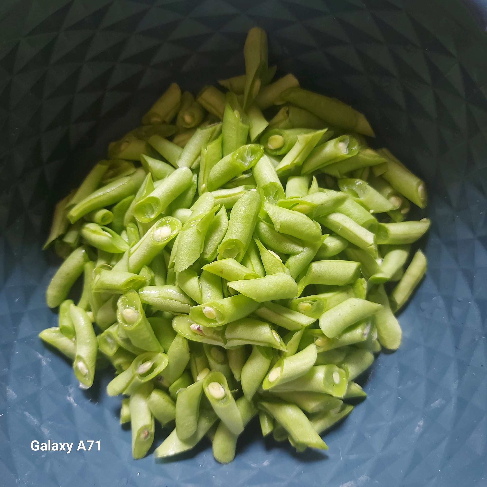 A pile of chopped green beans is in a dark blue plastic bowl with a textured surface. The beans are cut into small, uneven pieces, with some showing their white interiors and seeds. The bowl is filled with the beans, which are densely packed. In the bottom left corner of the image, the text "Galaxy A71" is visible. The background of the bowl has a diamond pattern. The lighting appears to be overhead, casting shadows on the beans and the bowl. The image is taken from a top-down perspective. There are no people present in the image. The location is unknown and cannot be determined from the image.