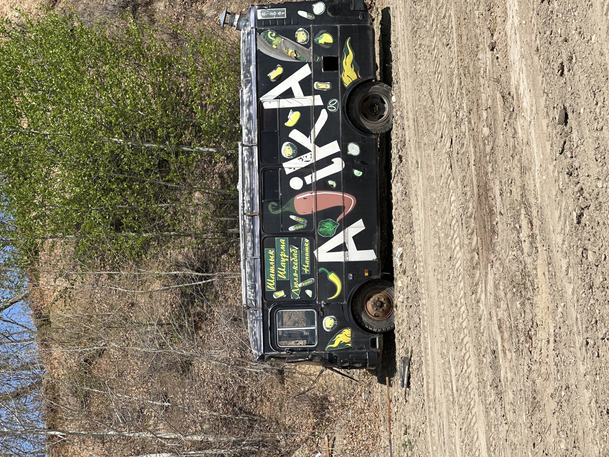 The image depicts a black food truck parked on a dirt road in what appears to be a wooded area. The truck has a large, white inscription on its side, along with images of peppers, herbs, and flames. There are also Cyrillic words, which translate to "Shashlik, Shawarma, Lyulya-kebab, Drinks". The location is identified by a sign on the truck as Chigiri, Russia.
