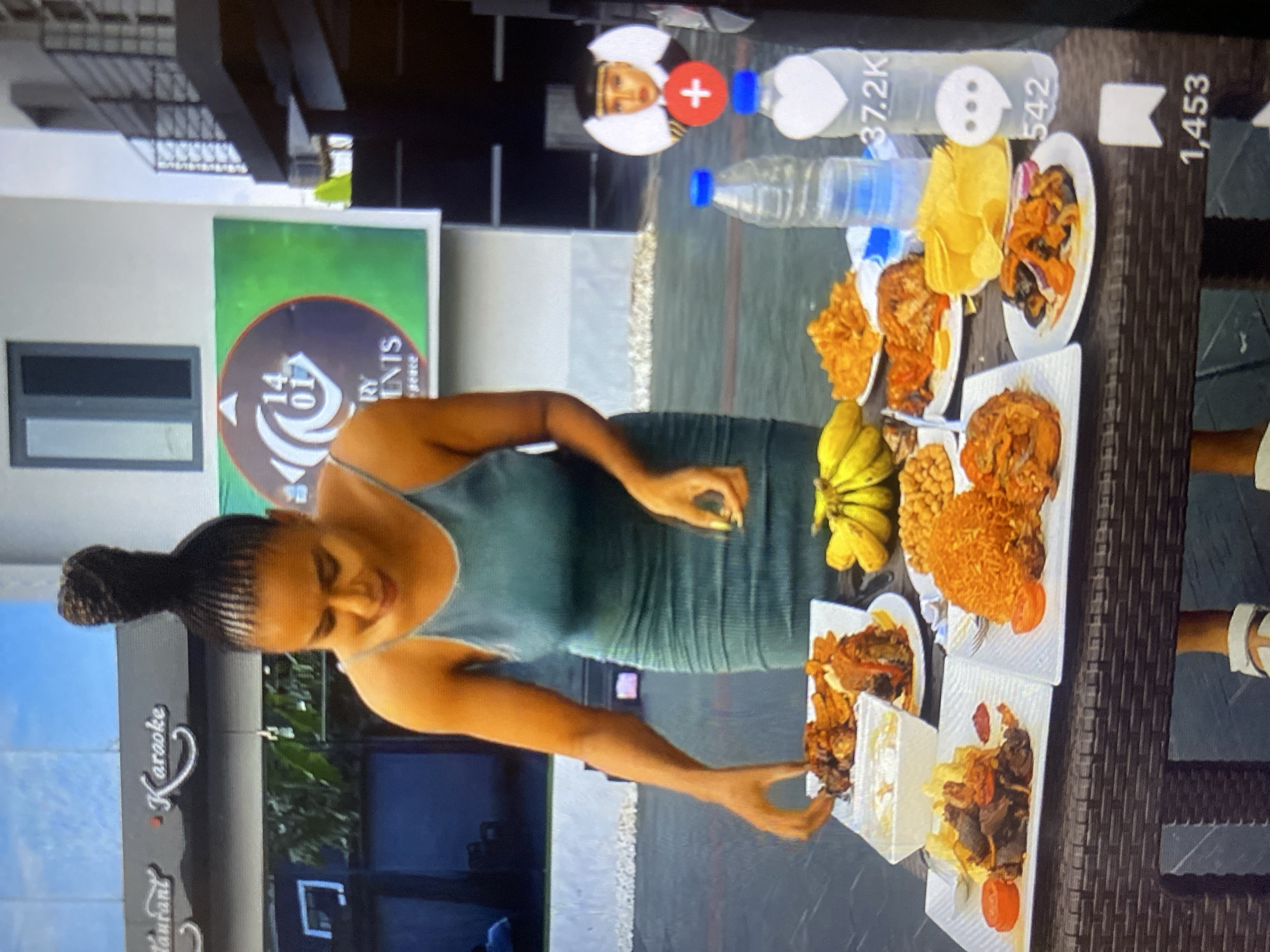 A woman with braided hair styled in a high bun, wearing a teal ribbed spaghetti strap dress, stands behind a dark brown outdoor table laden with multiple plates of food. Her head is bowed, and her left hand is positioned over a plate containing a meat dish and fried plantains.
On the table, there are several distinct dishes: a plate of jollof rice with chicken and a tomato slice, a plate of stewed meat with plantain fries and a tomato slice, a white bowl with a creamy liquid or semi-solid dish, a plate of various roasted or fried items including meat and a red sauce, a plate of yellow potato chips, a plate of dark stewed vegetables with onions, a bunch of yellow bananas, and a small bowl of chickpeas or groundnuts. Two plastic bottles of water, one clear and one with a white heart decal, are also on the table.
The background features a building with a black sign displaying "Restaurant" (partially obscured) and "Karaoke" in white script. Further back on a lighter-colored building, a large rectangular sign with a green background and a circular logo is visible. The logo contains "14 01" and partially readable text "RY ENIS" followed by "peace". A dark metal balcony railing is present on the far right. The scene is located in the city of Howayi, Nigeria. Digital overlays, indicative of a social media platform, are present on the right side of the image, showing a profile icon, a red plus sign, a heart icon with "37.2K", a speech bubble icon with "542", and a bookmark icon with "1,453".