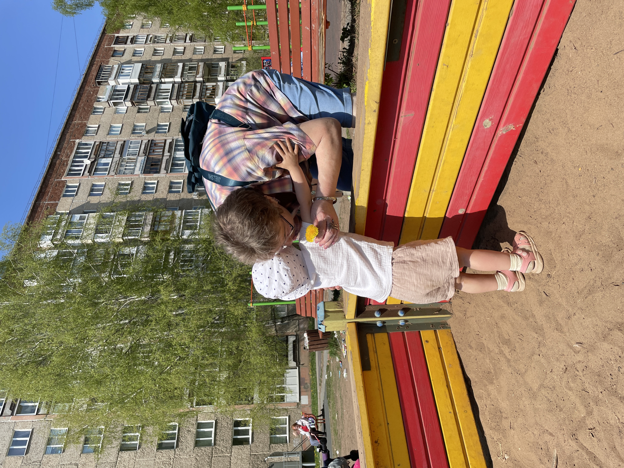 A child in a white hat, shirt, and pink skirt stands next to a brightly painted red and yellow structure in a sandy playground. The child holds a yellow flower. An adult sits on the structure, leaning over to interact with the child. A backpack rests against the adult’s back. Behind them, a multi-story apartment building is visible, along with trees and other playground equipment. The scene is in Izhevsk, Russia.