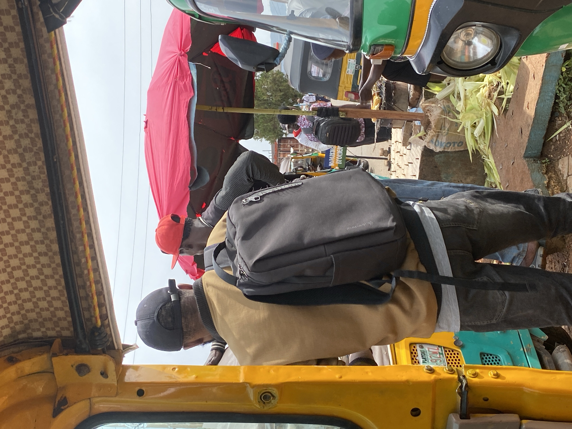 The image depicts a busy scene in Jos, Nigeria, likely a roadside market or transport hub. The primary subjects are people, appearing to be passengers or vendors, interacting with several colorful auto-rickshaws, also known as 'keke'.
One auto-rickshaw, bright yellow, is prominently featured in the foreground, its interior visible, adorned with a patterned upholstery. A person wearing a black cap and a tan jacket with a dark backpack is seated in this vehicle, looking downwards. Another person, wearing an orange cap, is visible in the background, partially obscured by a large, bright pink umbrella.
Behind the auto-rickshaws, other individuals are seen, some potentially vendors, with goods such as corn husks visible near a green auto-rickshaw. The overall atmosphere suggests a bustling, active environment, characteristic of a market or transportation point. The sky appears overcast, hinting at a potentially warm and humid day.
Notable details include the various vehicles and their vibrant colors, the presence of luggage like backpacks, and the assortment of people engaged in their daily activities. The ground appears to be dirt or gravel, typical of many roadside locations in Nigeria.
A license plate is visible on one of the vehicles, displaying "PLATEAU 924 LZ". This indicates the vehicle is registered in the Plateau State of Nigeria, reinforcing the location context of Jos, which is its capital.
The scene conveys a sense of everyday life and commerce, with people going about their business amidst the lively backdrop of the auto-rickshaws and market activities.