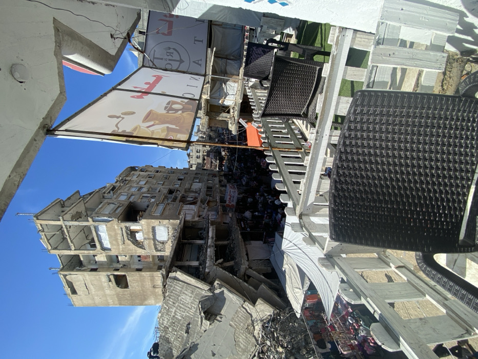 The image depicts an elevated view of a heavily damaged urban street in Khan Yunis, Palestinian Territory, under a clear blue sky. The perspective is from a balcony or terrace featuring white picket railings, artificial green turf, and dark wicker-style chairs. A large, tilted banner displaying coffee-related graphics and text, including "TRC" and "TANIC," hangs partially overhead.
Below, the street level is densely populated with numerous individuals engaged in market activity. Multiple market stalls, covered by white and orange canopies, line the street, displaying merchandise, possibly clothing.
On the left side of the street, a large, multi-story concrete building is extensively damaged, with significant portions of its structure collapsed, exposing internal floors, support beams, and rebar. Adjacent to and behind it, another multi-story building also shows severe damage, including numerous broken or missing windows and visible structural compromise. Further along the street, additional damaged and partially collapsed buildings are visible amidst debris.