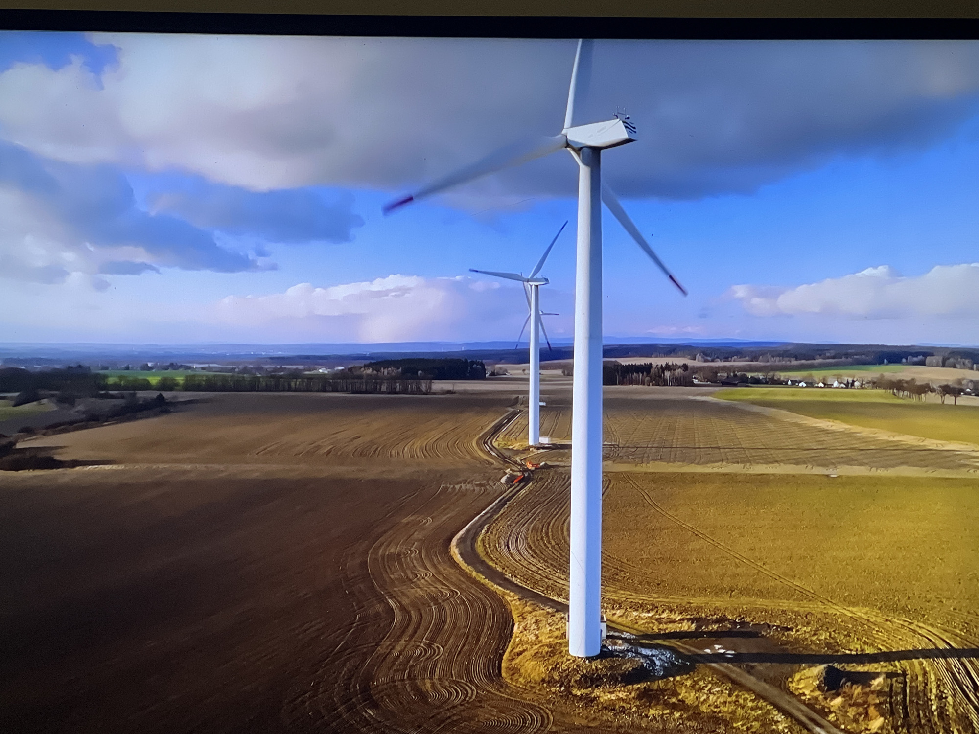 The image shows an aerial view of a wind farm under a cloudy sky. The foreground is dominated by three tall, white wind turbines, with their blades blurred due to motion. A winding dirt road, marked by tire tracks, leads between the turbines and into a field of tilled soil. In the background, a rural landscape unfolds with fields of varying colors, trees, and distant buildings. The sky is a mix of blue and gray clouds, with shafts of sunlight breaking through. There are no people or animals visible in the image. The location is not in Sofia, Bulgaria; it is a rural landscape.