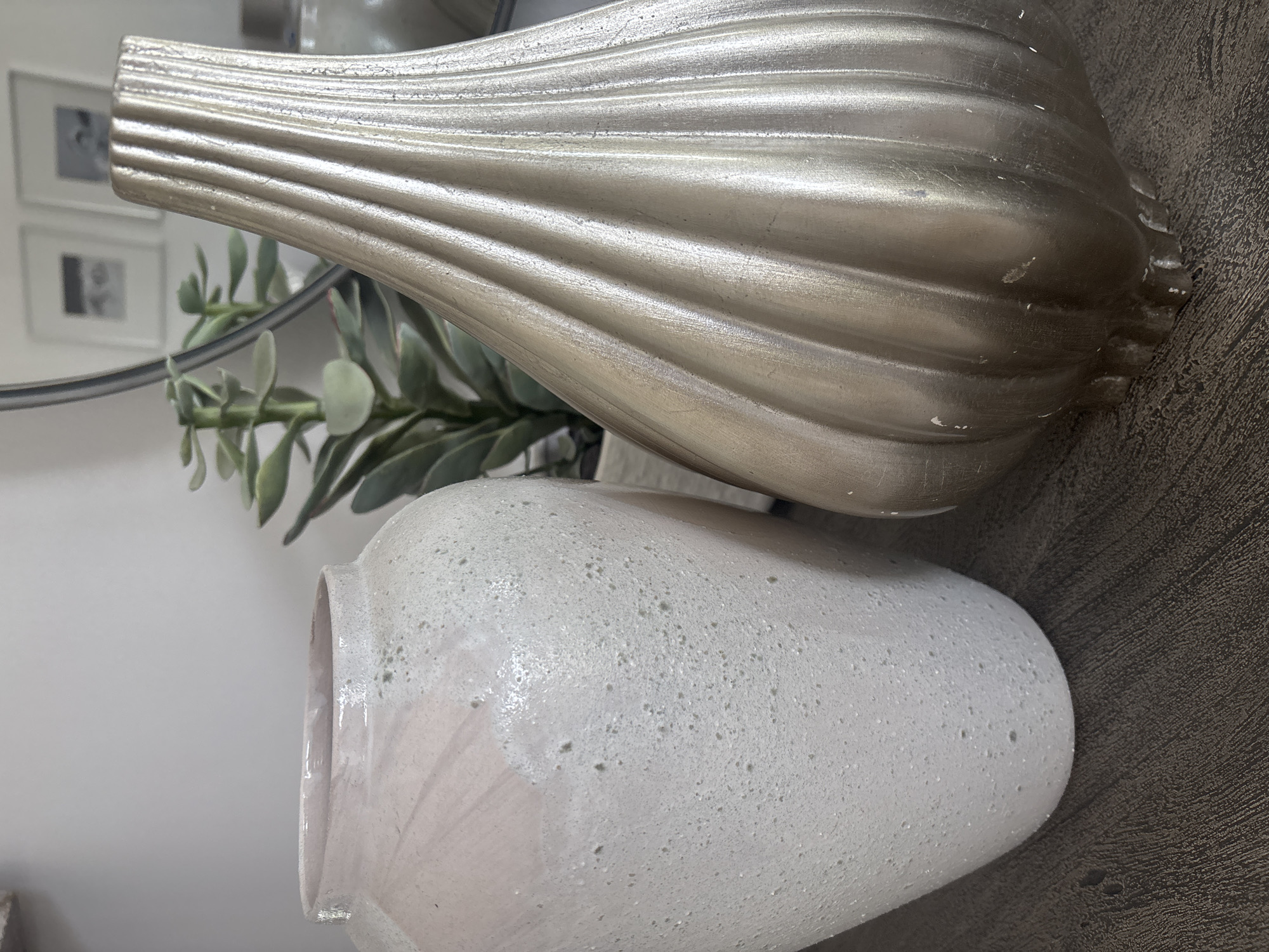 Two decorative vases are displayed on a dark wood surface. The vase on the left is a pale pink, speckled ceramic with a matte finish. The vase on the right is a metallic gold, with vertical ribbed detailing. Behind the vases, a green artificial succulent plant is visible, and a circular mirror reflects two framed pictures on a wall. The scene appears to be an indoor display, likely in a home or retail setting. There is no indication that this scene is located in Dubai, United Arab Emirates.