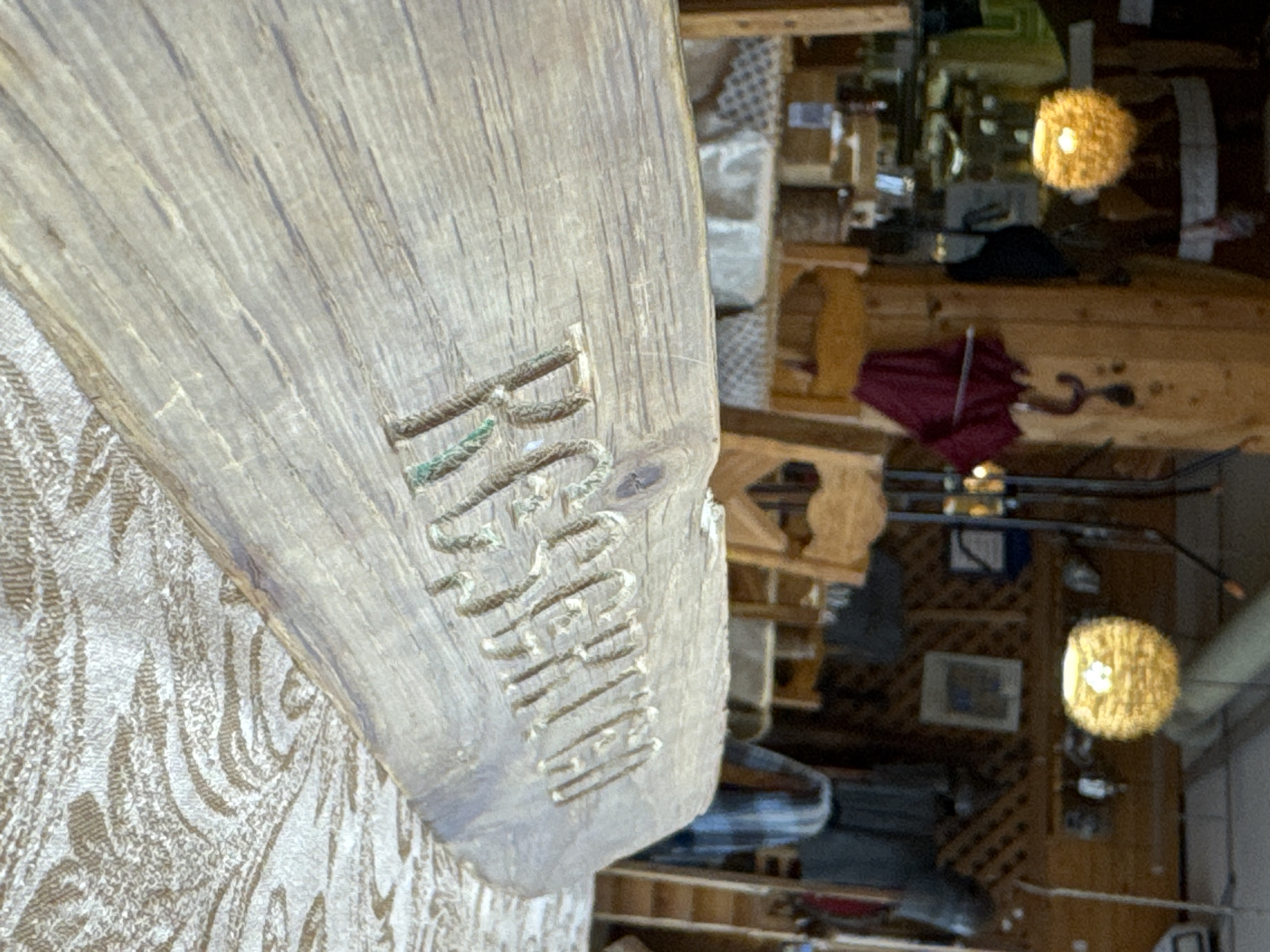 The image displays an indoor scene, likely a rustic shop, workshop, or a themed room, viewed at a tilted angle. The foreground is dominated by a thick, rough-hewn wooden plank, carved with the word "RUZHERO". The letters are deeply cut into the wood and show traces of a greenish-blue substance, possibly an inlay, paint, or natural aging like moss. The plank rests on a light-colored fabric with an ornate, swirling pattern.
In the background, the setting appears to be warm and dimly lit by artificial light sources. Two distinct, round pendant lamps, seemingly made of woven material like rattan, cast a warm glow. Various wooden structures, including shelves, a counter, and lattice work on a wall, are visible. Among the objects in the background are a red folded umbrella with a curved wooden handle, a dark cap or hat, and a pair of skis or similar equipment leaning against a wall. The overall impression is one of a cozy, possibly artisanal, environment. No people are present, and no specific activity is taking place; the scene is static. The location context indicates this is in Ново-Никольское, Russia.