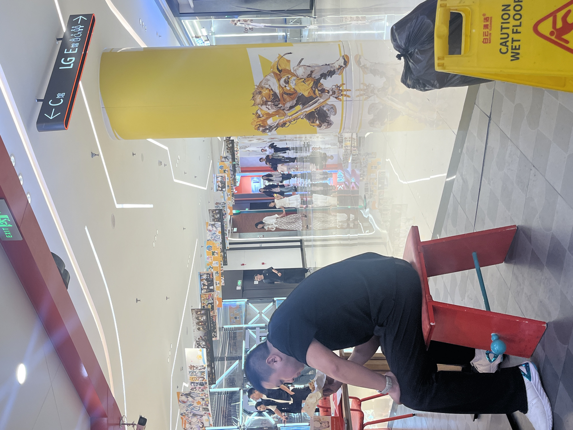 In Yuzhong District, China, a man in a black t-shirt and black pants sits on a red stool in a brightly lit indoor environment, likely a shopping mall. He is holding and eating food wrapped in white paper. His left hand is near his mouth, and his right hand is on the food. The stool has a teal colored bar across the front and two blue decorative spheres attached. He wears white athletic shoes with a blue and white design on the heel. In the background, a large yellow pillar is adorned with a graphic of a character in yellow armor holding a weapon. To the right of the pillar, a black trash bag stands next to a yellow "CAUTION WET FLOOR" sign. Further in the background, several people are walking, and anime-style posters are displayed on a ceiling-mounted track. A directional sign with Chinese characters and icons for restrooms is visible overhead. The floor is highly reflective, showing the ceiling lights and surrounding environment.