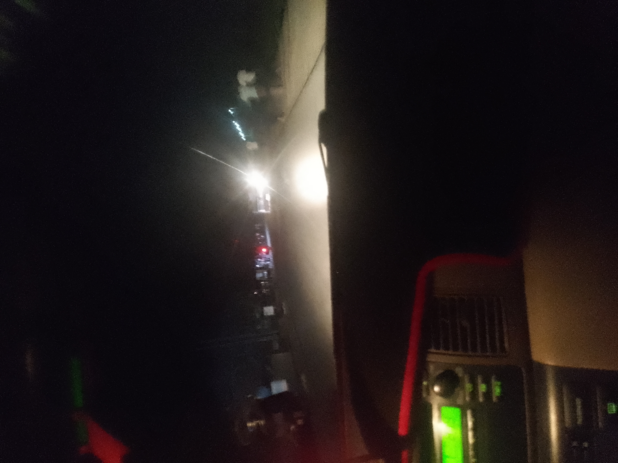 The image shows a view from inside a vehicle at night, looking out at a road with traffic. The road is illuminated by headlights and streetlights, creating a blurred effect due to the movement of the vehicle. Several vehicles are visible in the distance, appearing as streaks of light. The interior of the vehicle is dark, with some equipment visible on the right side, including what appears to be a vent and some control panels with illuminated green indicators. The image is grainy and lacks sharp detail, suggesting it was taken in low-light conditions. The context provided suggests this scene is in Jalingo, Nigeria, indicating an urban nighttime road environment.