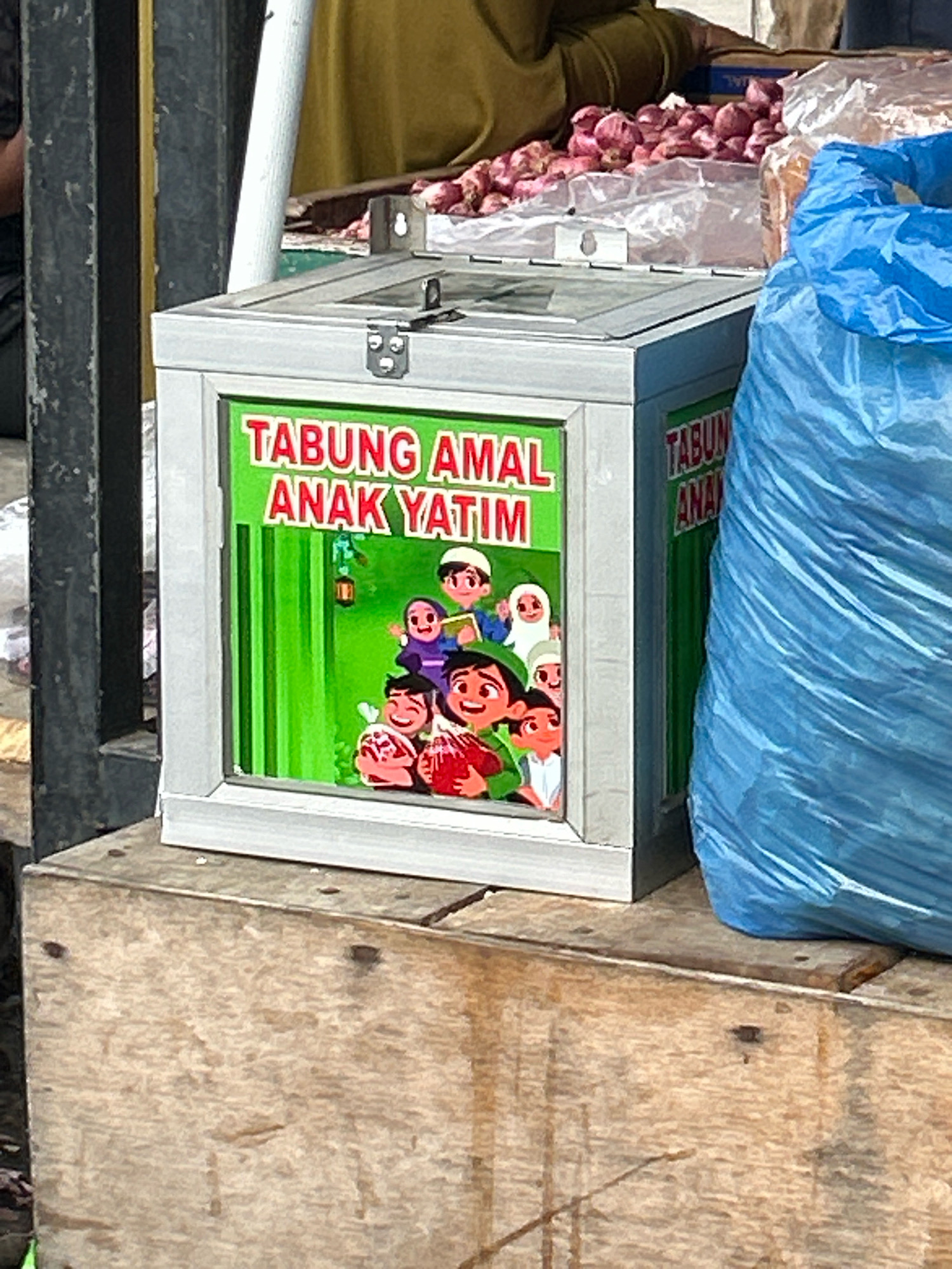 A collection box labeled "TABUNG AMAL ANAK YATIM" (Orphan Charity Box) in red letters against a green background with cartoon depictions of children is displayed prominently in the foreground. The box is made of metal with a hinged lid and is situated on a wooden surface. To the right of the collection box, a large, crinkled blue plastic bag is visible. In the background, a pile of red onions is present, suggesting a marketplace or food stall setting. The overall scene appears to be outdoors, possibly in a busy street or market area. The context suggests this image was taken in Banda Aceh, Indonesia, given the Indonesian language on the collection box.
