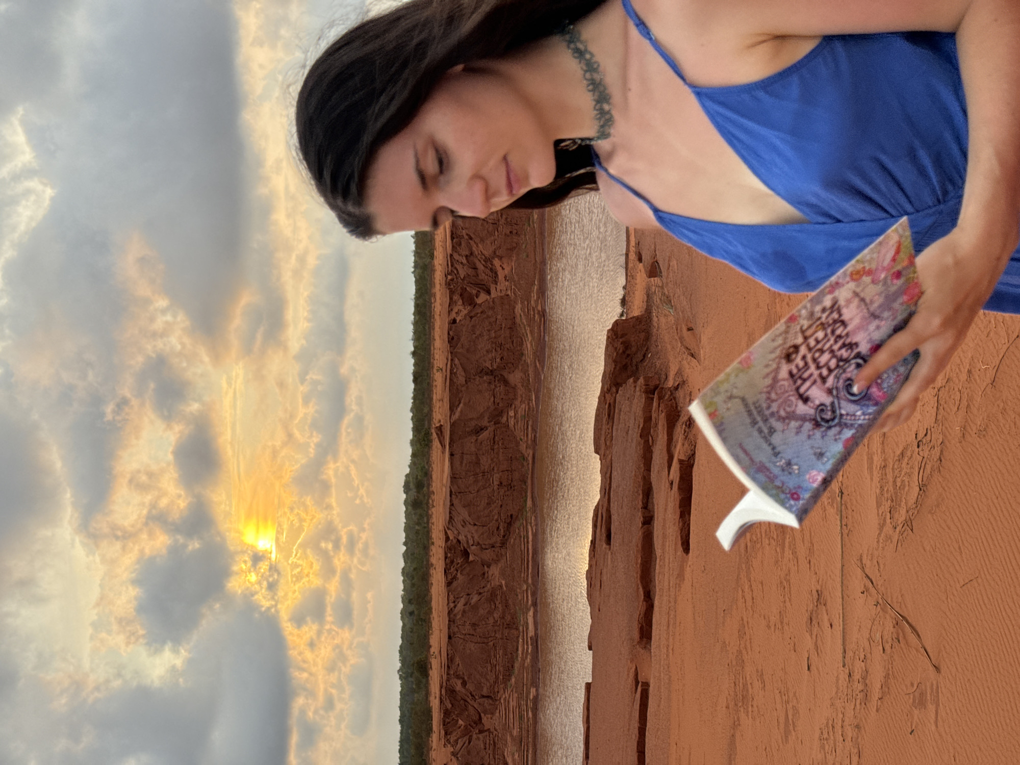 A young woman in a blue sundress stands outside in Mui Ne, Vietnam, holding an open copy of "The Secret Garden". The terrain behind her consists of red and brown rock formations with some foliage along the top edge. Water reflects the light in the distance. The sky is cloudy with the sun breaking through.