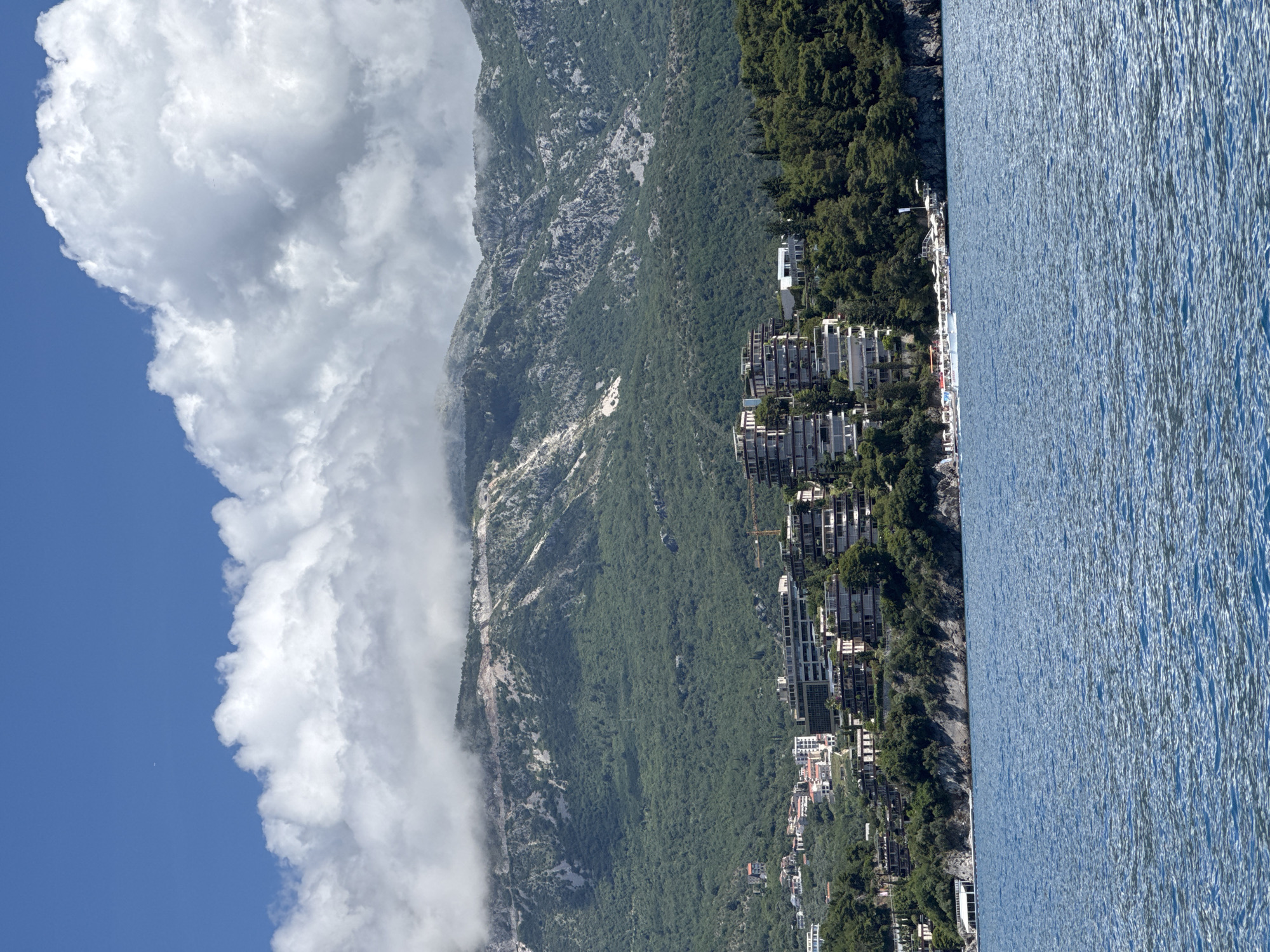 This striking landscape image captures a picturesque coastal scene, identified as Budva, Montenegro, under a bright daytime sky. The photograph is oriented vertically, showing a vast expanse of calm, blue water on the right, meeting a rugged and heavily vegetated coastline on the left.
Along the immediate coastline and ascending the lower slopes, numerous modern buildings are visible. These structures, characterized by their contemporary design with multiple levels and balconies, appear to be hotels, resorts, or residential complexes, suggesting a vibrant tourist or residential area. They are interspersed among dense green trees, blending into the natural environment. Several smaller structures and what might be a pier or dock are also visible at the water's edge.
Further inland, the landscape rises dramatically into large, forested mountains. The lower and middle sections of these mountains are covered in lush green vegetation, while higher peaks reveal rocky outcrops and more sparse foliage. A significant cloud formation is draped across the middle and upper parts of the mountains, creating a soft, misty effect and partially obscuring the highest terrain.
The sky above is a clear, vibrant blue, contrasting beautifully with expansive, bright white cumulus clouds that fill a large portion of the upper left. The sunlight appears strong, illuminating the water and the buildings, indicating a sunny day. No people or specific activities are distinctly visible, but the developed coastline implies human presence and resort life. The image conveys a sense of serene beauty, combining natural grandeur with modern coastal development. No visible text is present.