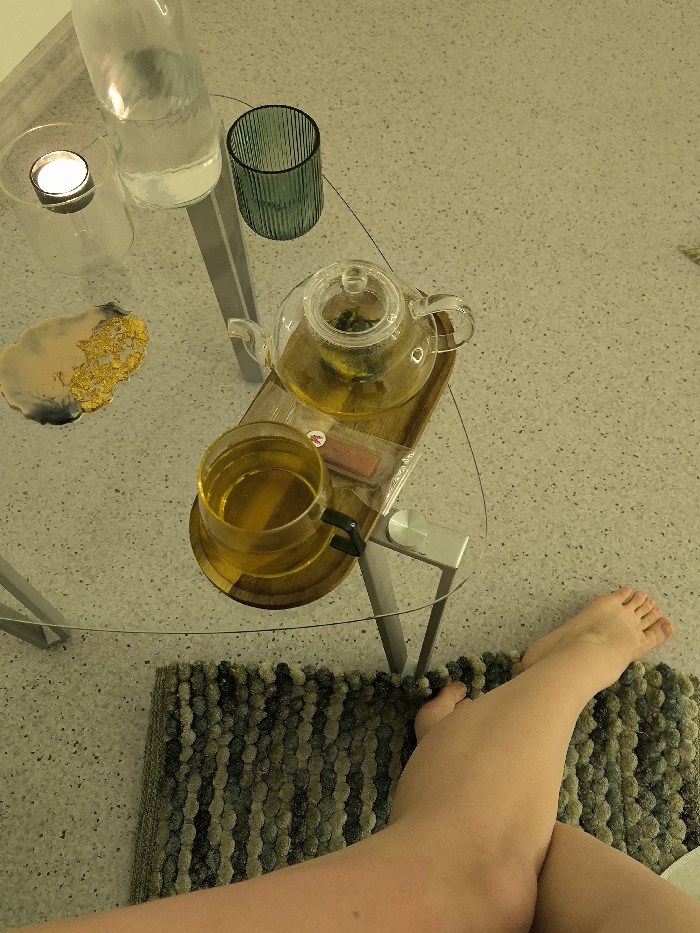 An overhead view reveals an indoor scene in Vinnytsia, Ukraine, featuring a clear glass triangular table with metal legs positioned on a light-colored, speckled floor. On the table, from left to right, are a clear glass bottle containing water, a clear glass votive holder with a lit tealight candle, and a decorative coaster with beige, gold, and dark blue elements. To the right, a dark green ribbed glass is present. In the center of the table, a small wooden rectangular tray holds a clear glass teapot filled with amber liquid and plant matter, indicative of brewed tea. Next to the teapot on the tray is a small, clear package containing a light-colored rectangular item. A clear glass mug with a dark handle, also containing amber liquid, rests directly on the table surface. A person's bare legs are visible in the lower right foreground, crossed at the ankles, with their feet resting on a textured, looped-pile rug exhibiting shades of green, grey, and black.