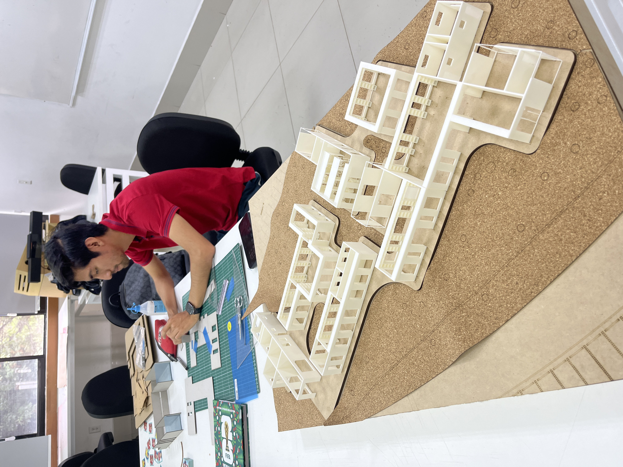 A person in a red shirt is working on a model architecture project in a well-lit indoor space. The model, consisting of white structures, is laid out on a corkboard base. Adjacent to the model on a table are tools such as a cutting mat, ruler, scissors, glue, and various building materials. The project is in San Andrés Cholula, Mexico. The room appears to be a workshop or studio setting.