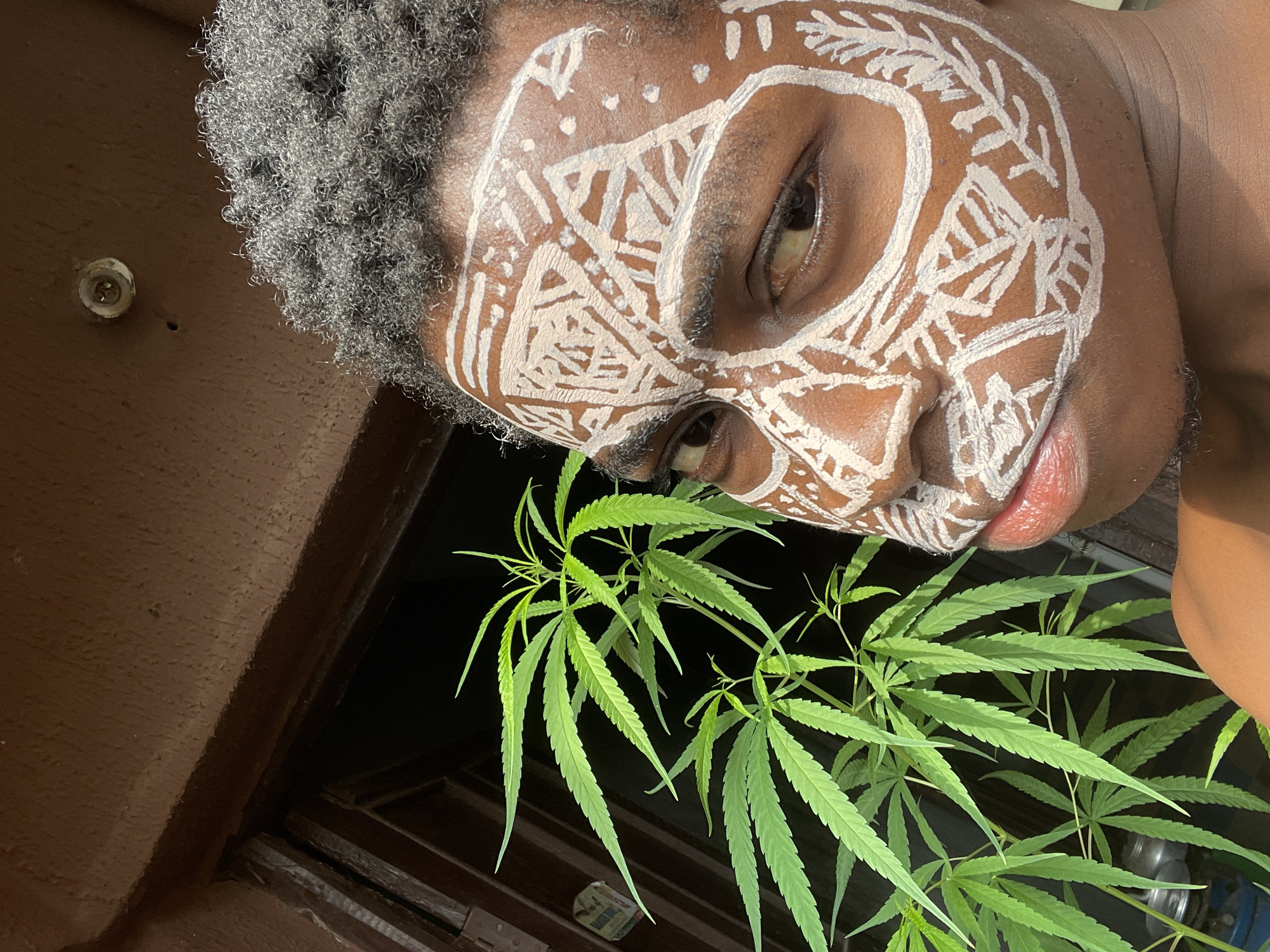 A close-up, tilted shot shows a person with brown skin and short, gray, tightly curled hair. The person's face is painted with white designs resembling a mask. Below the face are green marijuana plants. The background appears to be a dark window frame and a brown wall. The location is Nkpor, Nigeria.