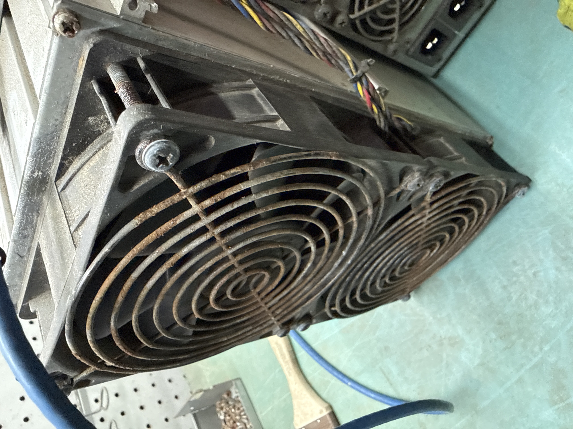 The image shows a close-up, low-angle view of two large, dusty computer cooling fans mounted on industrial equipment, possibly part of a cryptocurrency mining rig. The fans have rusty grilles and are secured with visible screws. A bundle of colored wires is tied with black zip ties, emerging from the side of the equipment. A blue electrical cable snakes across the foreground, and a paintbrush with a wooden handle lies on a light green, textured surface in the background. The overall scene suggests a workshop or industrial setting. No specific location in Indiantown, United States of America, is discernible from the image itself.
