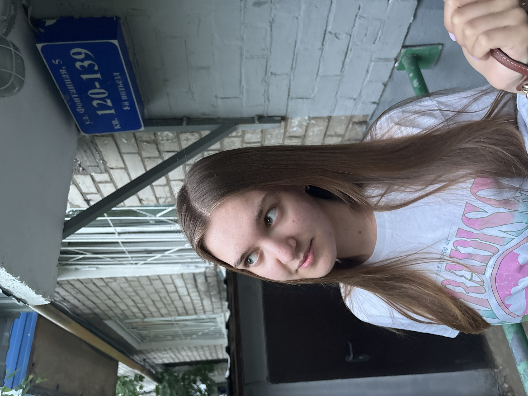 A young woman stands outdoors next to a building in Perm, Russia. The street sign attached to the building reads "ul. Fontannaya, 5, kv. 120-139, 8-i pod'yezd." The woman is wearing a white t-shirt with a colorful graphic design. The building has a brick wall and a metal awning structure.