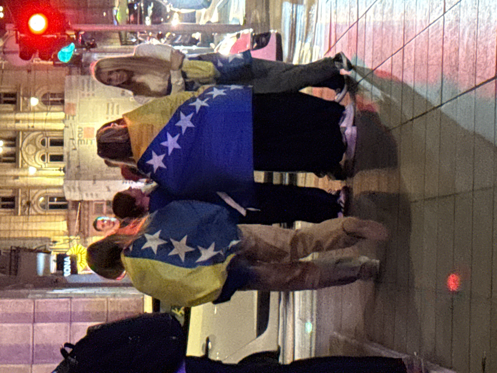 A group of people are standing on a sidewalk at night, with a traffic light visible in the background. The individuals are wearing casual clothing, and two of them are draped with flags. The flag on the left appears to be the flag of Bosnia and Herzegovina, featuring a blue field with a yellow diagonal band and white stars. The flag on the right is also partially visible and seems to share similar colors and star patterns. The surroundings suggest an urban environment with buildings and streetlights. The ground is paved with tiles, and reflections of lights are visible on the wet surface, indicating recent rain or damp conditions. The overall scene is dimly lit, with artificial lighting contributing to the atmosphere. The presence of the Bosnian flag suggests a potential gathering or event related to Bosnia and Herzegovina, possibly in Sarajevo given the flag's prominence.