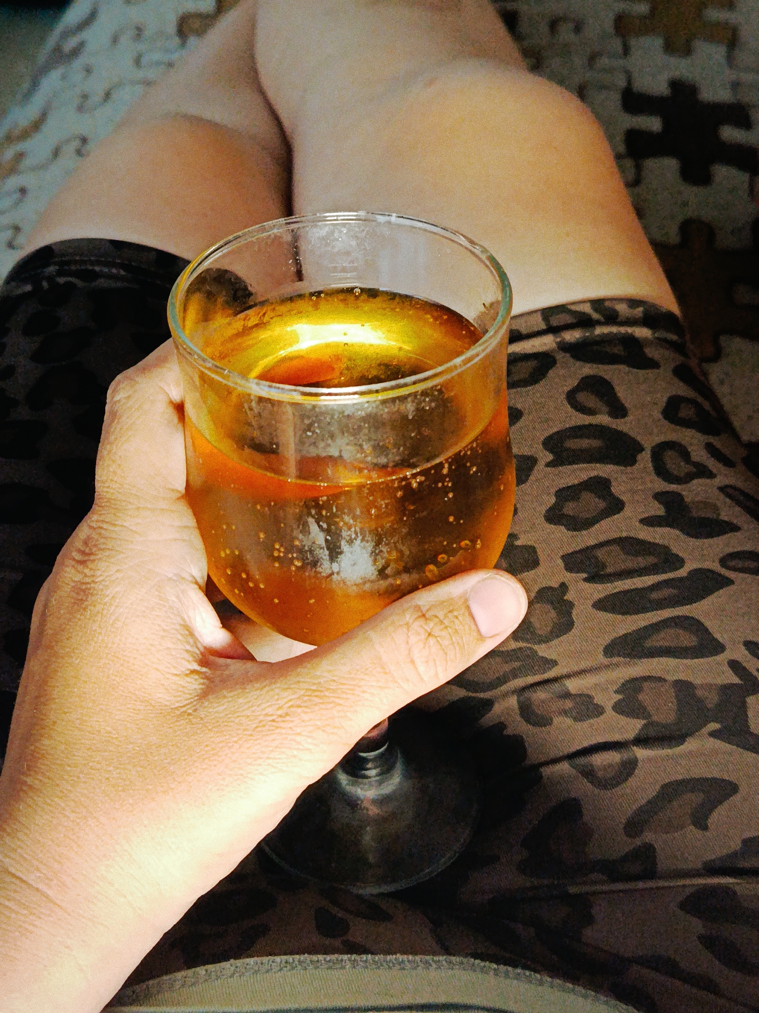 A person is holding a glass of amber-colored liquid, possibly a beverage, in their right hand. The glass has a short stem and a wide, rounded bowl. The liquid contains small bubbles. The person is sitting with their legs crossed. They are wearing clothing with a leopard print pattern. The background includes a carpet or rug with a pattern of interlocking puzzle pieces in muted colors. There is no information to support that this image was taken in Фоки, Russia.