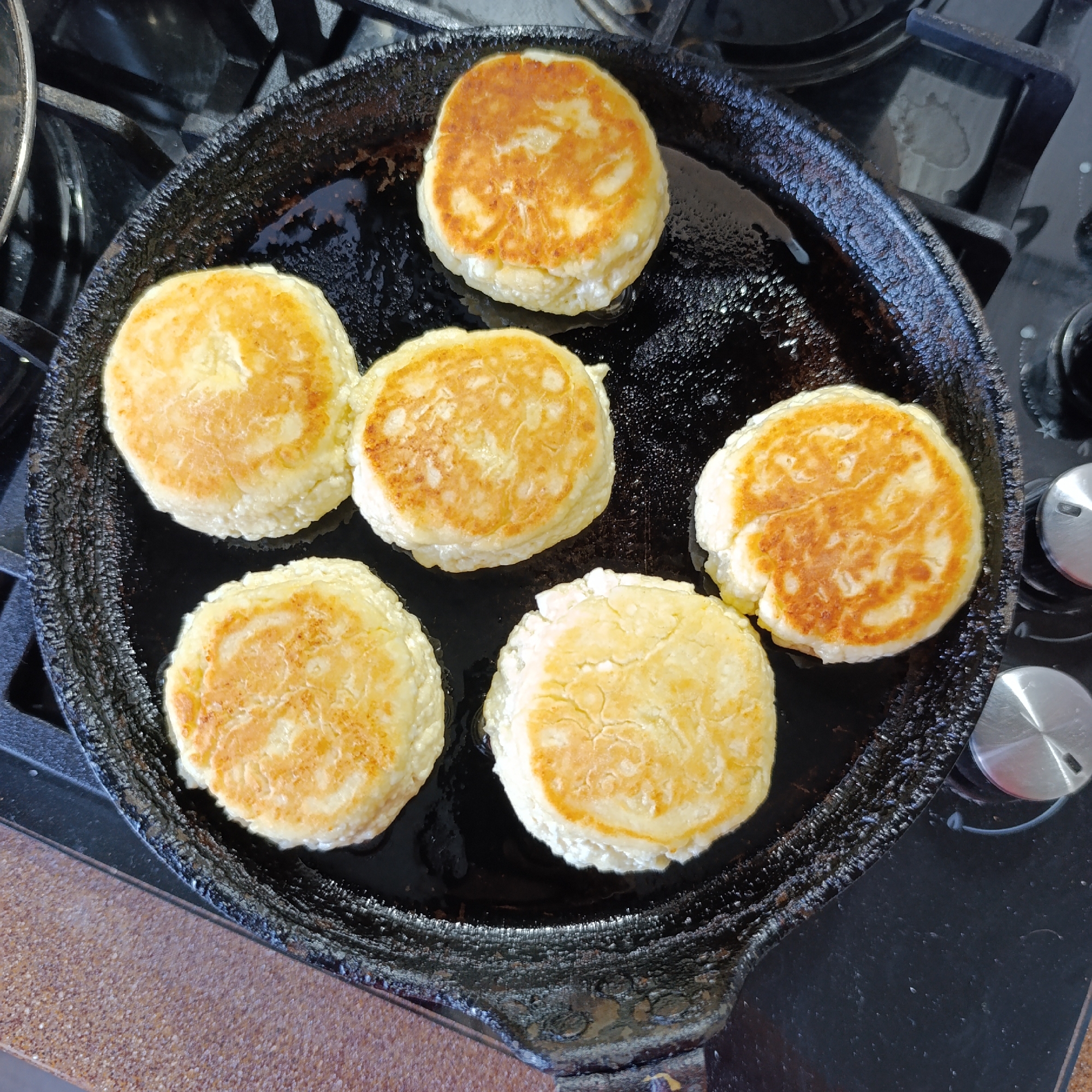 The image depicts a black cast iron skillet filled with six circular, golden-brown pancakes. The skillet rests on a black stovetop, with burner controls visible on the right. The pancakes are evenly spaced within the pan, surrounded by cooking oil. The countertops near the stove are brown. The image was taken in Марковский, Russia.