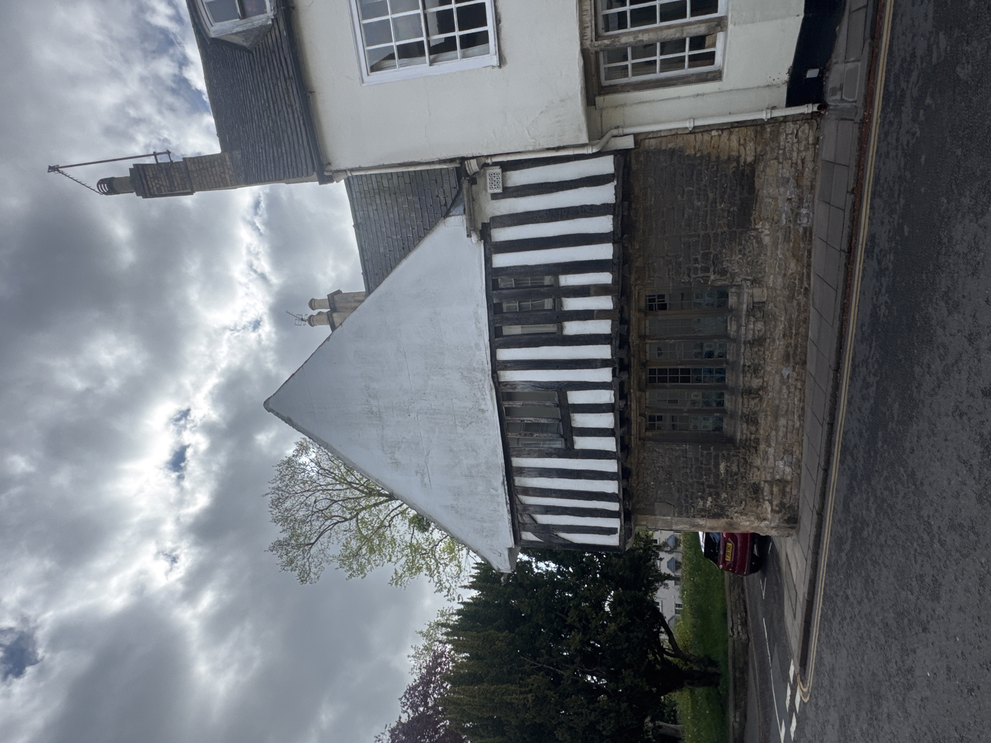 The original image, rotated 90 degrees clockwise, presents a street view of historic architecture under a cloudy daytime sky in what appears to be South Kesteven, United Kingdom. The central building exhibits a blend of architectural styles: an upper section rendered in white with white-framed, multi-paned windows; a distinctive middle section showcasing traditional half-timbering with black beams and white infill, containing smaller windows; and a lower section built from rugged stone or brickwork, featuring larger, possibly leaded, windows.
A prominent brick chimney, topped with an antenna or aerial, extends from the roofline on the left. Further to the left, another building with a dark-tiled roof and a white upper facade is partially visible. The scene includes a paved road with typical UK double yellow lines and white arrow road markings, adjacent to a sidewalk. A red car is partially parked by the road, revealing the beginning of a UK license plate: "FJ XXK". The setting is softened by several trees, displaying both fresh light green and darker, denser foliage, beneath a sky filled with grey clouds. No specific human activity or strong emotions are evident, conveying a quiet, architectural street scene.