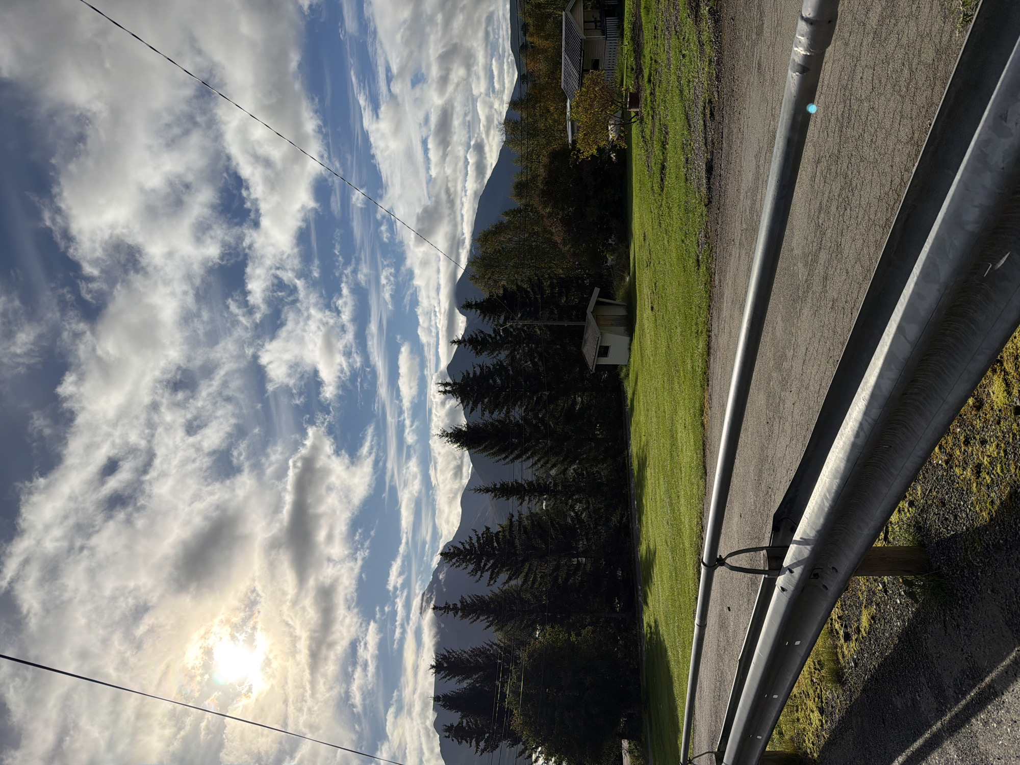 The image depicts a rural scene with a mountainous backdrop, likely in Storsteinnes, Norway, under a partly cloudy sky with the sun partially obscured. In the foreground, a metal guardrail lines a gravel road. Beyond the road is a vibrant green lawn, leading to a line of dark green coniferous trees. Behind the trees, several buildings are visible, including a small, white shed and a larger house with a dark roof. The mountains in the distance are shrouded in clouds, with their peaks occasionally emerging. The overall impression is one of peaceful, natural beauty.