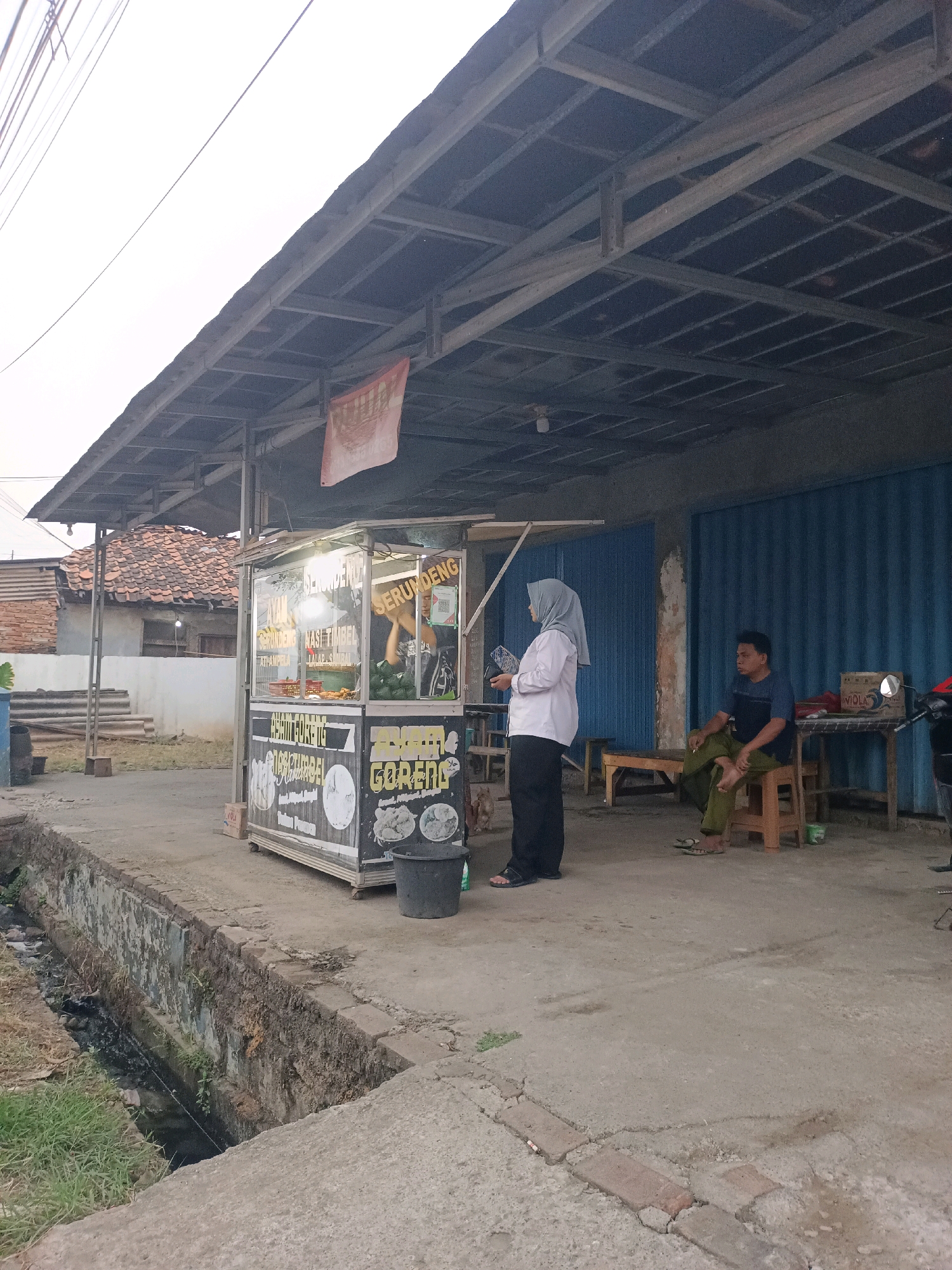 In Karawang, Indonesia, a street-side food stall operates under a corrugated metal awning. The stall is brightly lit, displaying a menu with Indonesian text and images of food items like "Ayam Goreng" (fried chicken) and "Serundeng" (fried coconut). A woman wearing a grey hijab and white shirt stands by the stall, holding a tablet or phone. To the right, a man in a dark t-shirt and green pants sits on a wooden stool, smoking a cigarette. Behind them, a blue roll-up door marks the entrance to a building. The environment shows a narrow concrete drainage ditch with murky water running alongside the paved area. In the background, traditional Indonesian houses with terracotta tile roofs are visible. A motorcycle is parked on the right side of the frame. The overall scene depicts a common street food vendor setting in a residential or commercial area.