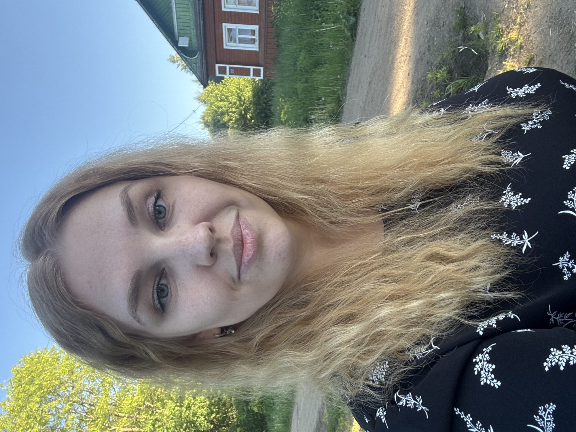 The image displays a young adult female with long, wavy blonde hair, light skin, and green eyes, looking directly at the viewer with a subtle smile. She has light freckles visible across her nose and cheeks. Her left ear features a small, silver-toned hoop earring with a dark, dangling charm. She is wearing a black long-sleeved top or dress patterned with numerous small, white, stylized floral or leafy designs.
The scene is outdoors on a clear, sunny day, indicated by the bright blue sky in the upper background. To the left, lush green deciduous trees are visible. In the background, on the right side of the frame, a traditional wooden house stands. It is painted a reddish-brown with contrasting white trim around its windows and corners, and features a steep, dark green gabled roof. The number "22" is visible on the side of the house. A dirt or gravel path runs across the mid-ground, bordered by tall green grass and various weeds. The location is identified as the city of Rozhdestveno, Russia.