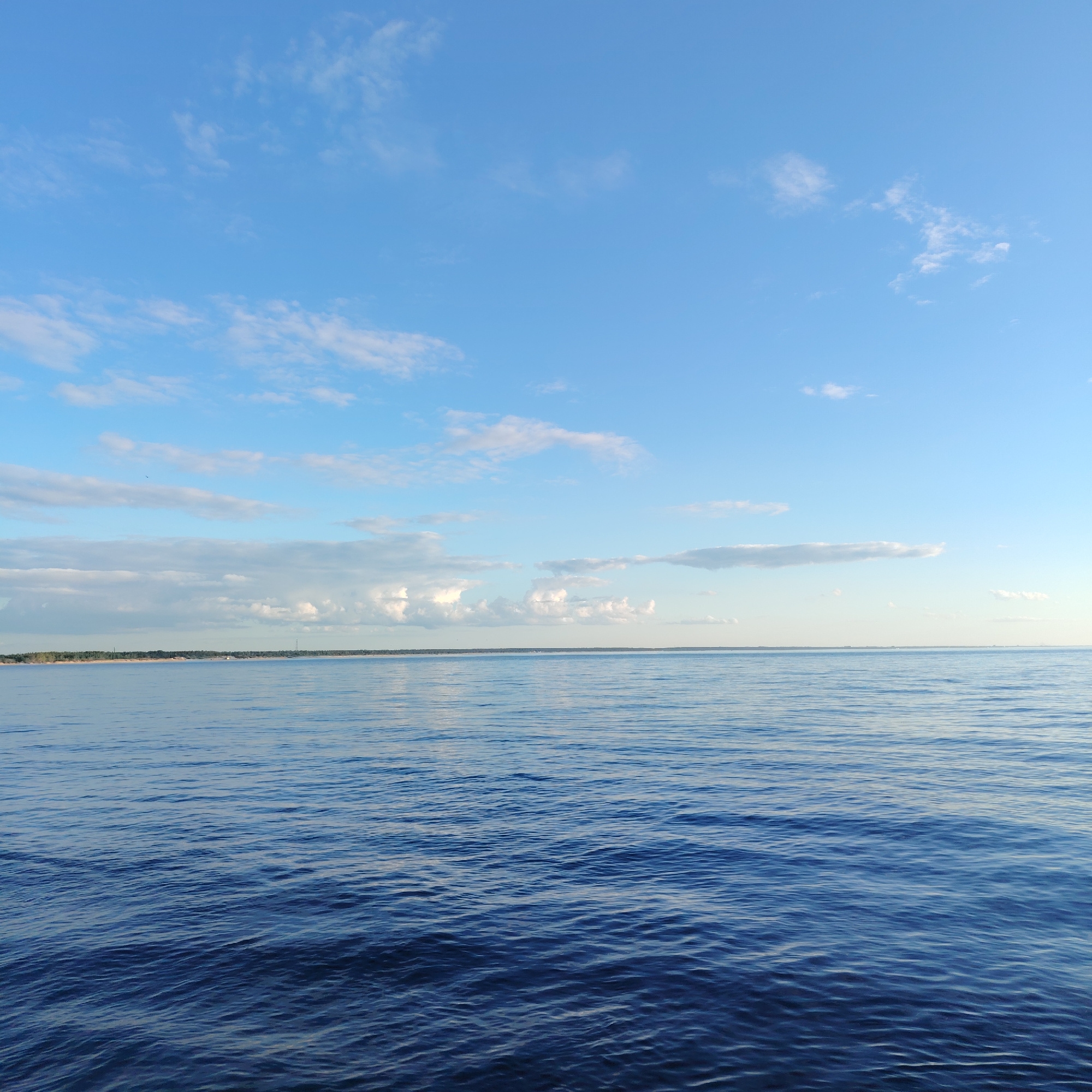 The image shows a body of water, likely the Baltic Sea, under a blue sky with scattered white clouds. A thin strip of land is visible on the horizon. Without additional landmarks or contextual information, pinpointing the exact location in Riga, Latvia is not possible. The scene depicts a calm and open environment with the sea taking up most of the frame.