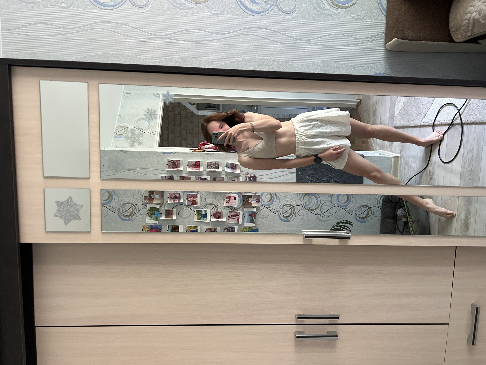 A young woman stands in a room with a large, mirrored wardrobe in Chaikovsky, Russia, and takes a selfie with her smartphone. She wears a light-colored top and shorts. The room's decor includes patterned wallpaper and a rug. The wardrobe is light-colored with drawers and mirror panels decorated with designs and photographs. An electrical cord is visible on the floor.