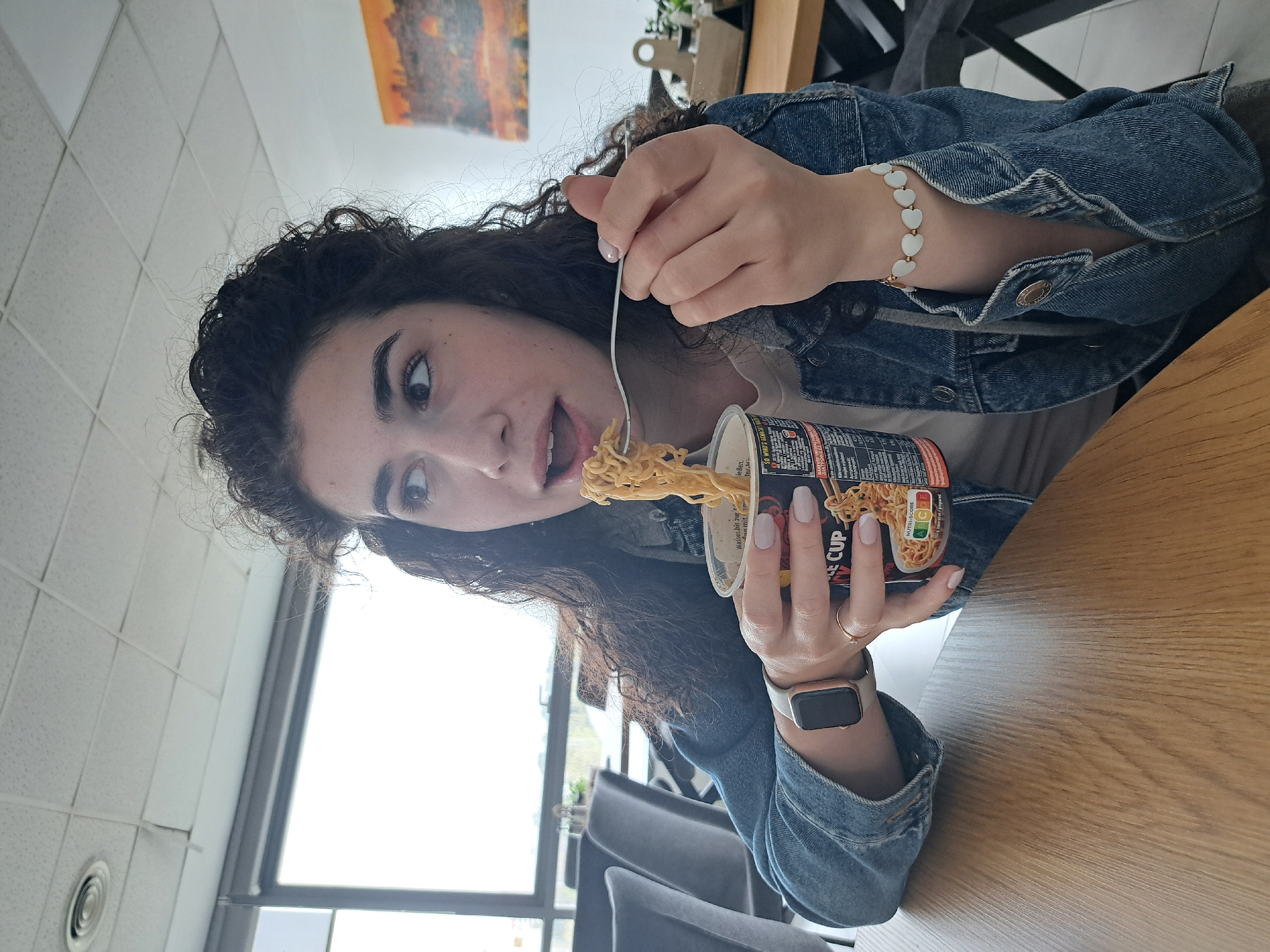 A young woman with dark, curly hair is sitting at a light-colored wooden table, eating noodles from a paper cup with a fork. She is wearing a denim jacket and a heart-shaped bracelet on her left wrist and an Apple Watch on her right wrist. She looks off to the left with a surprised expression, her mouth open as she eats.
The cup of noodles is labeled with nutritional information and the words "CUP" and "NUTRI-SCORE." In the background, there is a painting, chairs, and a window showing a cityscape with trees. The setting appears to be a casual indoor dining area. While there are some elements suggesting a location, the information needed to confirm its location in Sofia, Bulgaria is not available in this image.