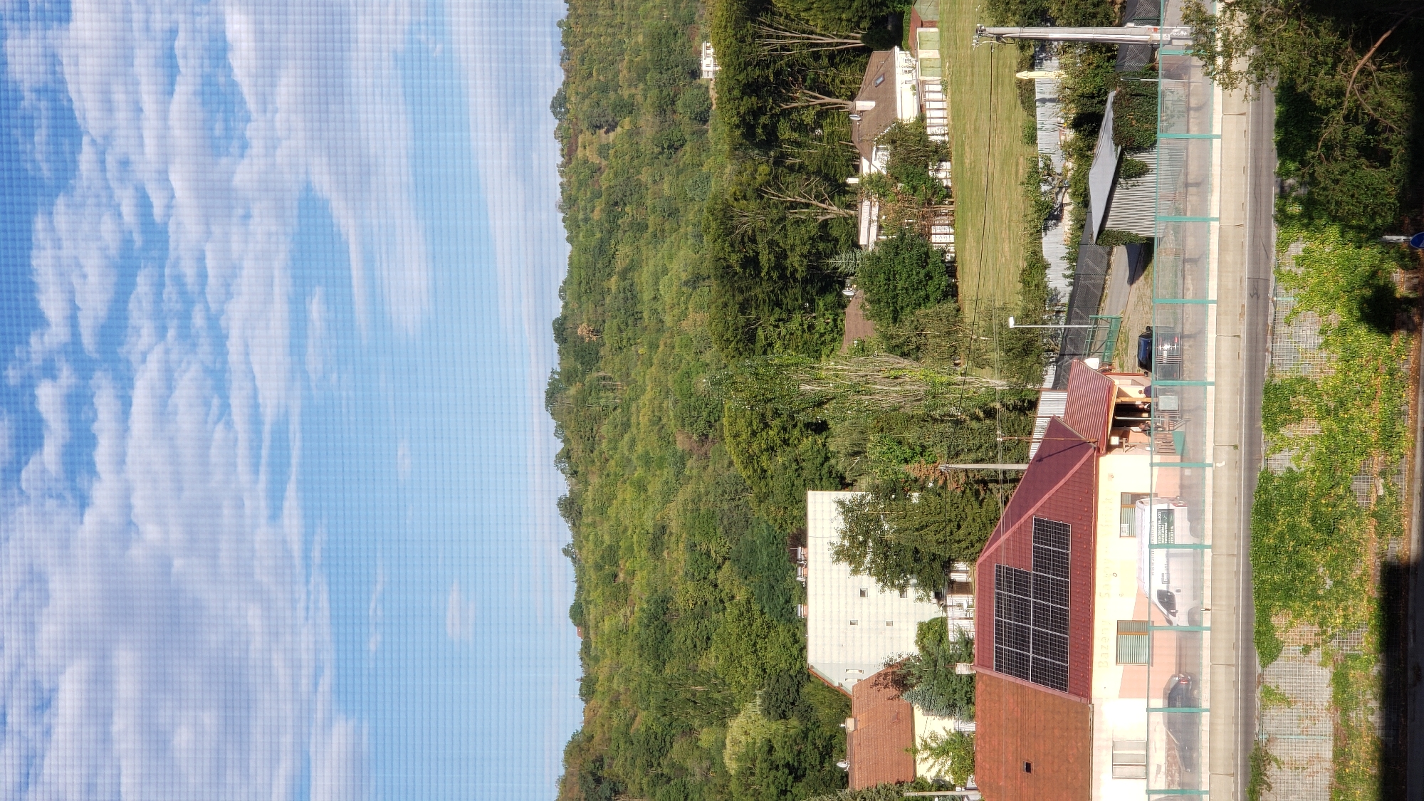 This is a daytime aerial view of a suburban area in Prešov, Slovakia, characterized by residential buildings, lush greenery, and a distant wooded hill. In the foreground, a two-story building with a red roof and solar panels is visible. Adjacent to it is a single-story structure with a covered outdoor seating area. A white van and a dark-colored car are parked on the street in front of these buildings, separated by a light green metal fence. Beyond the buildings, a gently sloping grassy field stretches towards a densely wooded hill that dominates the background. Several other houses with white fences are scattered across the landscape. The sky above is bright blue with scattered white clouds, and the overall scene suggests a peaceful, residential neighborhood on a clear day.
