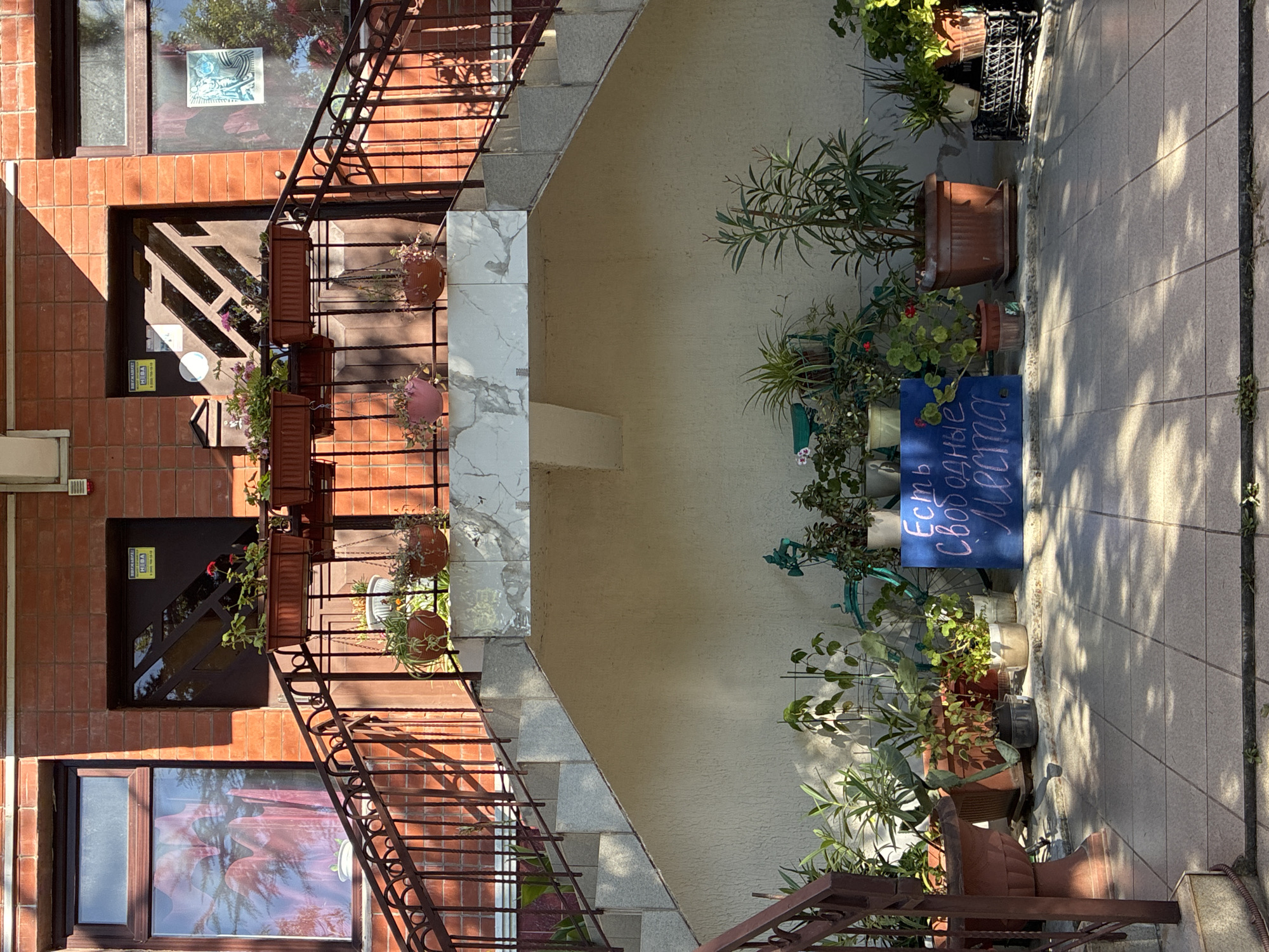 The image shows the exterior of a building in Sochi, Russia. A set of stairs with a reddish-brown metal railing leads up to two doors on the second floor. The building is constructed of red brick, and there are windows above the doors, one of which is partially obscured by a sheer pink curtain. Planters filled with plants and flowers are mounted on the railing.
The ground level features a tiled patio area with numerous potted plants and a bicycle. A blue sign with white lettering is placed on the patio, reading "Есть свободные места" (There are free spots). The overall environment suggests a residential or commercial building, possibly an apartment complex or a small business. The lighting indicates it is daytime, with shadows cast by the plants and the structure. There are no people visible in the image.