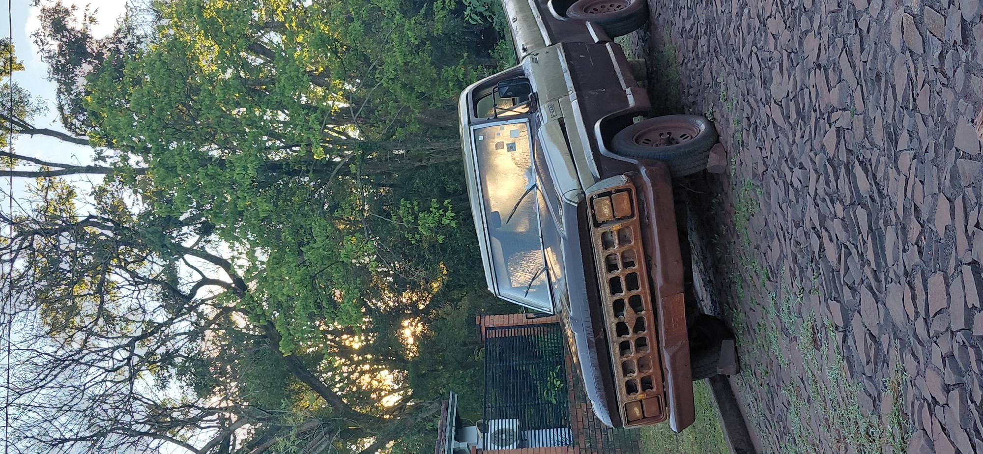 The image depicts a faded, two-toned pickup truck, possibly a Ford F-Series from the 1970s, parked on a cobblestone street in Encarnacion, Paraguay. The truck is a dark brown and tan color, with visible rust on its grille and front bumper. The windshield is reflecting the setting sun, which shines through a canopy of lush green trees in the background. A brick wall and fence are visible to the left of the truck, adjacent to a building. The scene is captured at an angle, with the truck positioned diagonally across the frame. No people are present in the image.