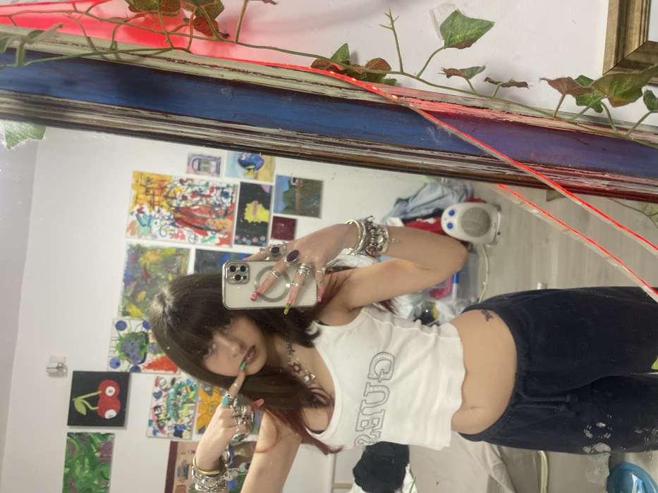 A young woman with bangs takes a mirror selfie, wearing a white tank top, dark pants, and numerous rings and bracelets. Her nails are painted in various colors. The mirror reflects a room with a white wall covered in colorful paintings, as well as a small white heater and other items. A vine and red string lights adorn the top of the mirror.