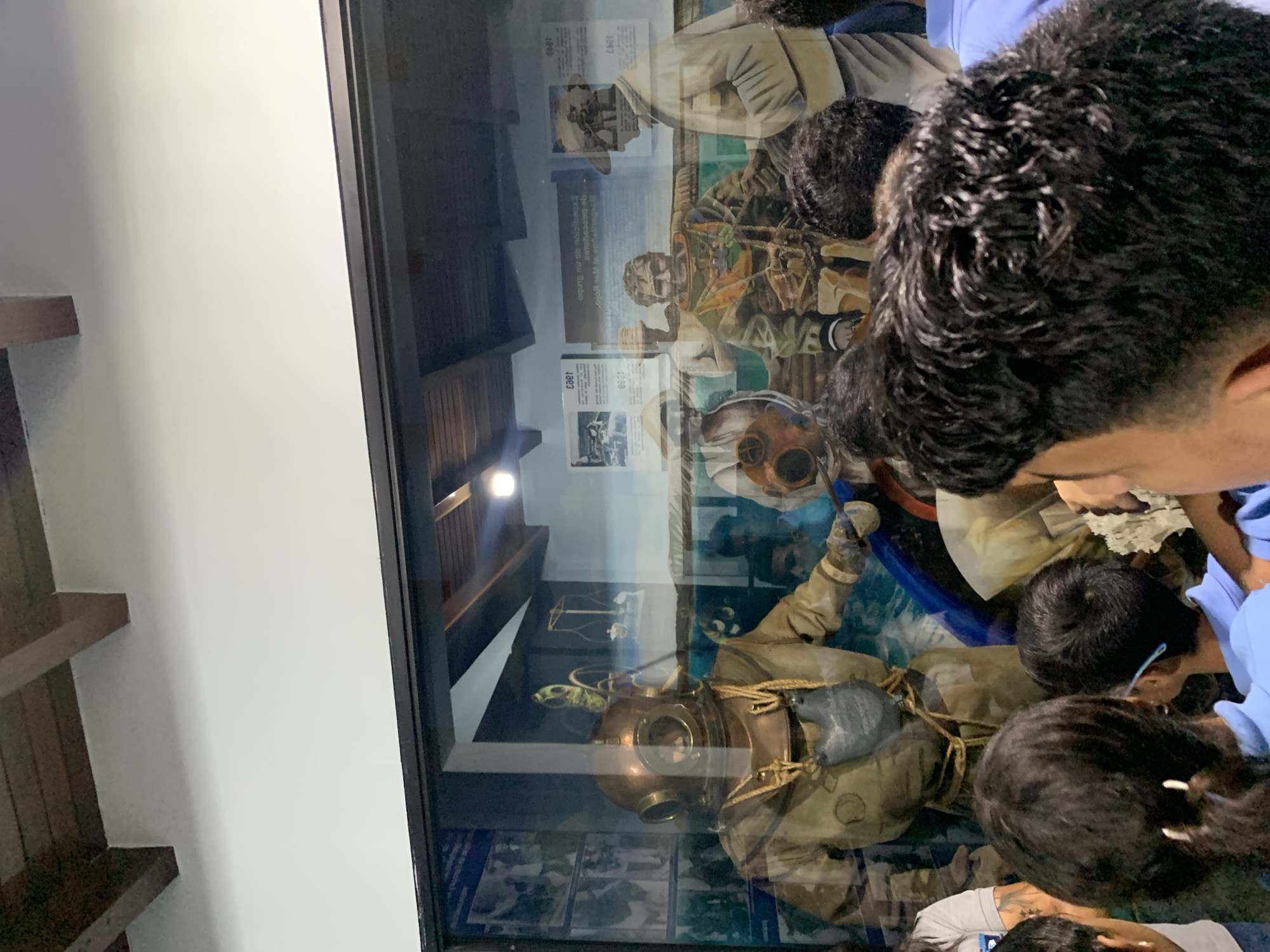 In Boca del Río, Venezuela, a group of people, primarily children with dark hair, are looking at a museum exhibit through a glass display case. The exhibit features mannequins dressed in vintage diving suits. One mannequin in the foreground is wearing a copper helmet and a tan suit with a gray pouch. Behind it, other mannequins depict divers in different stages of their gear, some with hats. The display includes historical photographs and text panels on the back wall, one of which appears to be a painted mural of divers and historical equipment, including a scale. The environment is dimly lit, with wooden beams visible on the ceiling. The overall scene captures a moment of learning and observation at a historical exhibit.