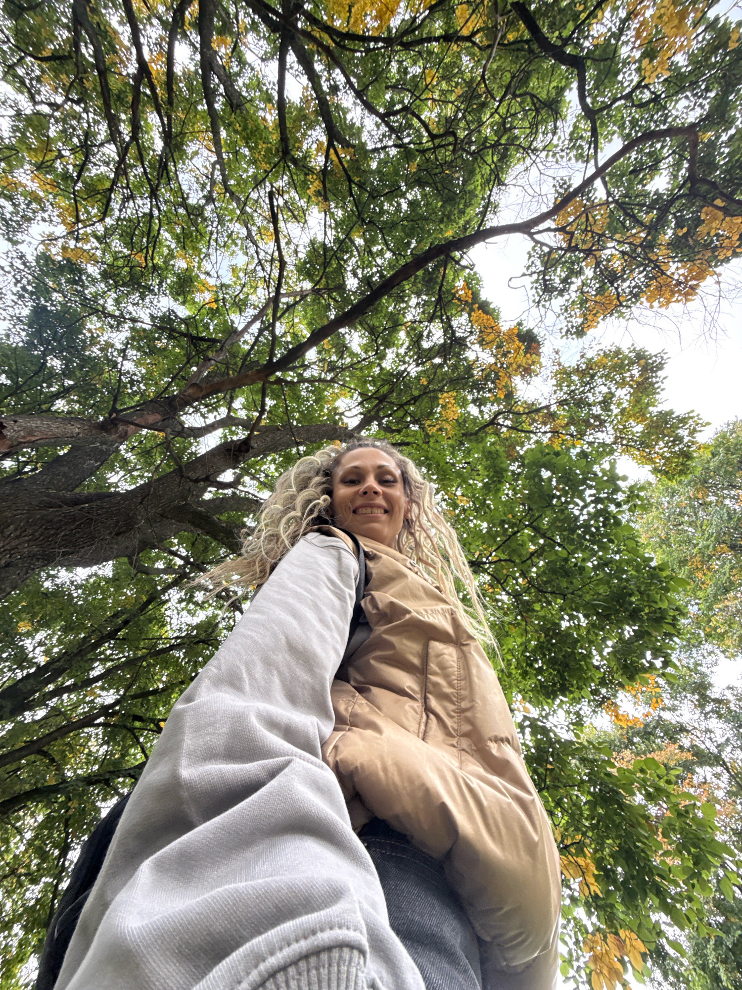 An eye-level, medium shot captures a young woman from a low-angle perspective beneath a canopy of trees. The woman is partially visible, with her head turned towards the camera and a smile on her face. She has long, curly blonde hair and is wearing a tan puffer vest over a light grey hoodie. Her arm is extended upwards, holding the camera. The background consists of tree branches with green and yellow leaves, indicating an autumn setting. The sky is visible through the gaps in the foliage. The overall scene suggests an outdoor environment, possibly a park or forest.