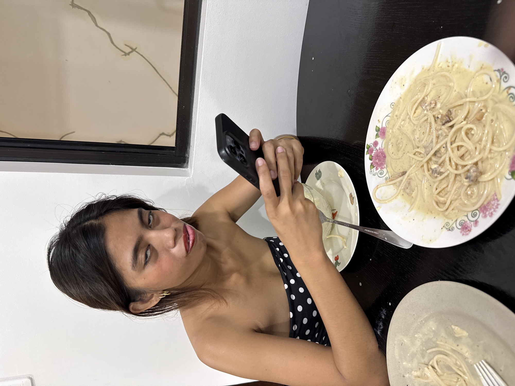 An adult female with medium-length dark hair, wearing a black and white polka dot strapless top, is seated at a dark wooden table, holding a smartphone in her hands and looking at it with a displeased expression. Her eyes are rolled back, and her mouth is pursed. In front of her, on the table, are two plates of spaghetti. One plate is partially eaten, while the other is fuller, with spaghetti and a creamy sauce. A fork is visible on the left plate, and another fork is in the bowl of spaghetti on the right. The background shows a white wall and a framed picture with a subtle pattern. The setting appears to be an indoor dining area, potentially in Davao City, Philippines, given the contextual information.