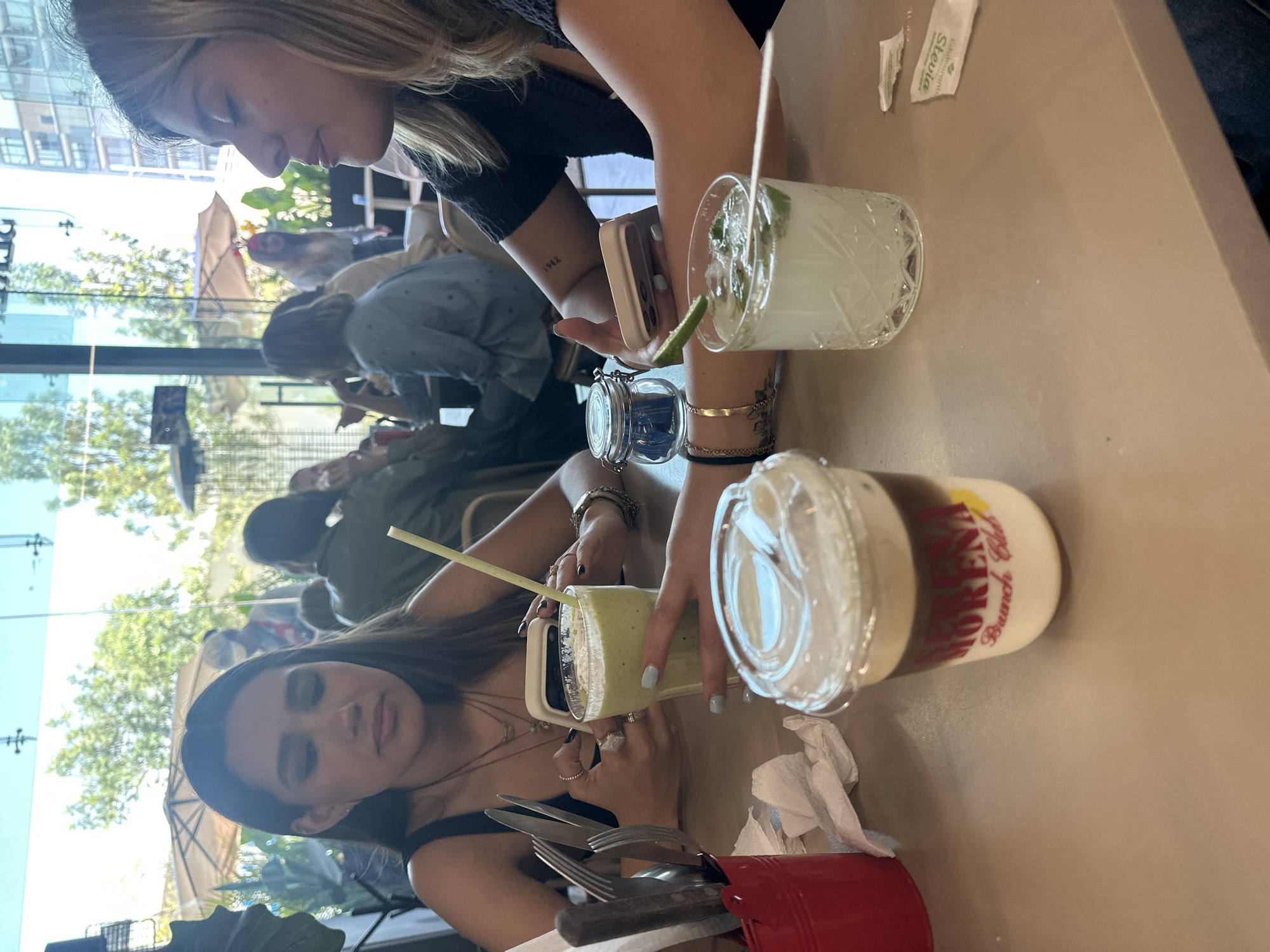 Two women are seated at an outdoor table at "Morena Brunch Club" in Puebla, Mexico, a popular restaurant. The location seems to be an open-air setting with a glass wall that allows views of outdoor scenery. One woman is using her smartphone, while the other appears to be sipping a green smoothie. Several drinks are present on the table, including a beverage in a clear, decorative glass and another in a plastic container. A red container of silverware is also visible. Other patrons can be seen through the glass wall.