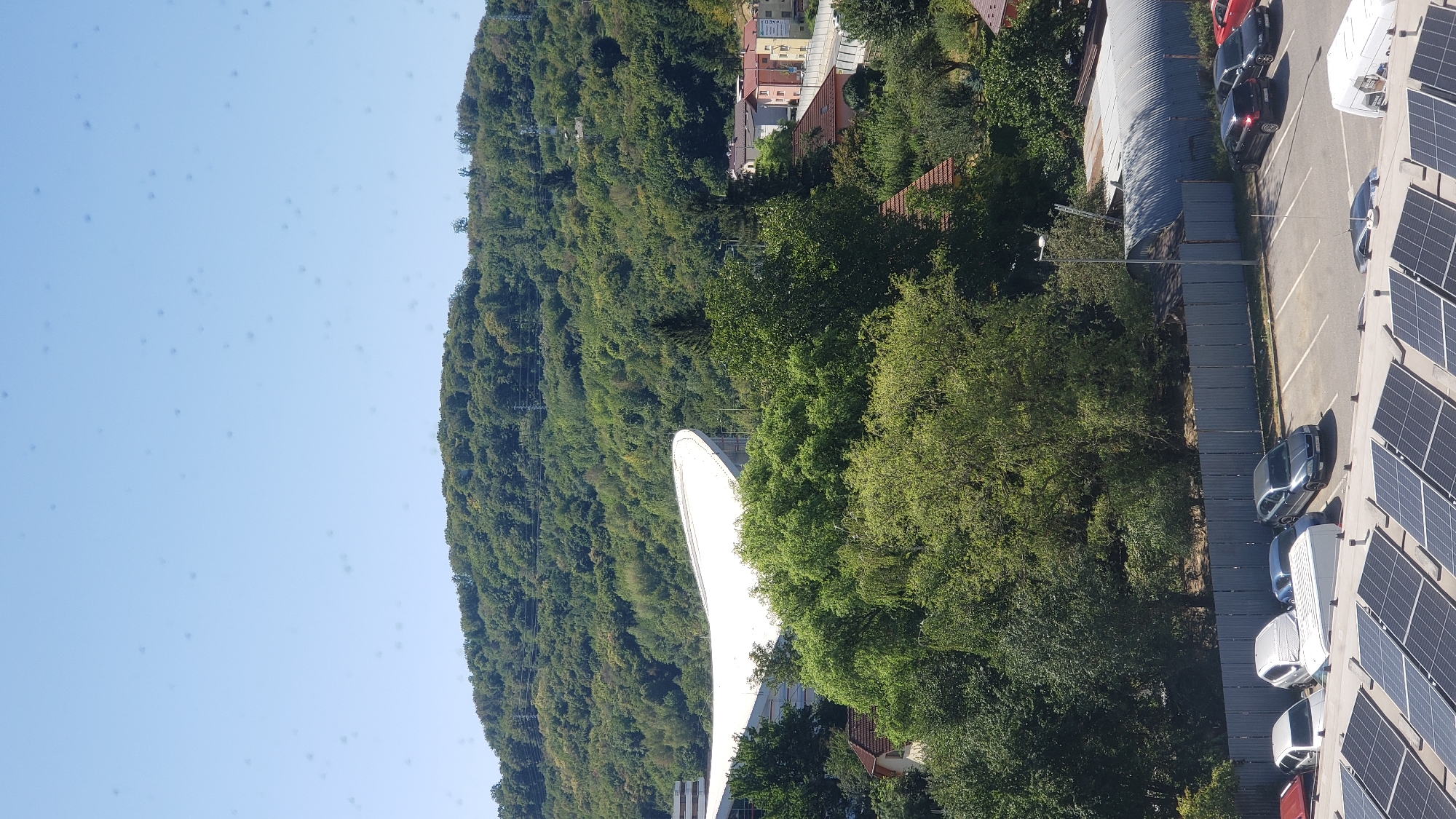 The image captures a view overlooking a suburban area on a clear day, with a dense green hill forming the backdrop. In the mid-ground, a large, white, curved structure, possibly a stadium or arena, is partially visible, nestled amongst lush trees. To the right of this structure, a cluster of residential buildings with red-tiled roofs and yellow and pink facades can be seen. A road runs alongside these houses. In the foreground, a parking lot is visible, with several cars parked, including dark sedans and a white van. A row of solar panels covers the roof of a building in the bottom of the frame. The scene appears to be located in the city of Prešov, Slovakia.
